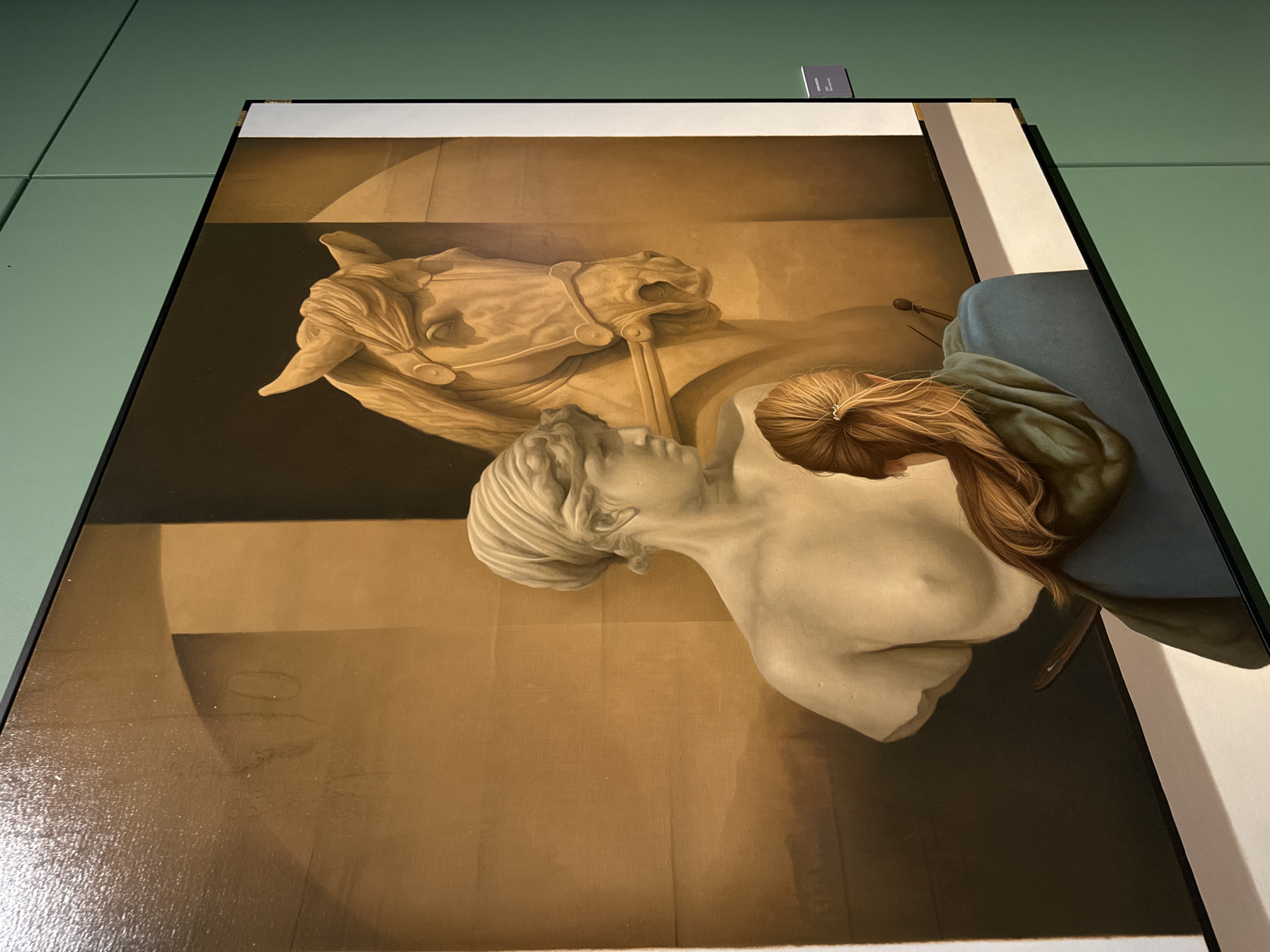 The image shows a framed painting of a horse head statue and a woman's bust. The painting is displayed on a wall in a gallery or museum. The location is in San Bernardino Tlaxcalancingo, Mexico. The subject of the painting is sculptural, with a classical style. The horse head has detailed features, including bridle decorations. The woman's bust is pale with realistic hair. The painting is predominantly in shades of brown, white, and gray. The frame is black. There is a small label on the top of the frame.