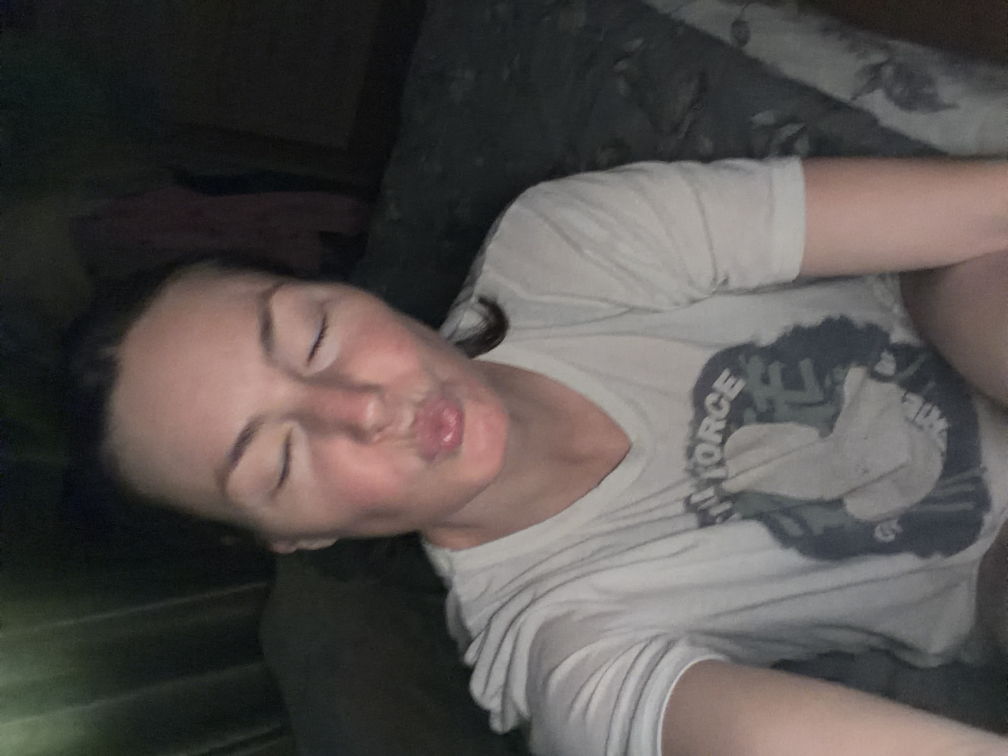 A person is lying on a dark-colored, possibly patterned, cushioned surface. The person is wearing a light-colored t-shirt with the words "AIR FORCE" visible. The person's eyes are closed and the mouth is pursed in a kiss-like gesture. The location in the city is Unknown, Unknown.