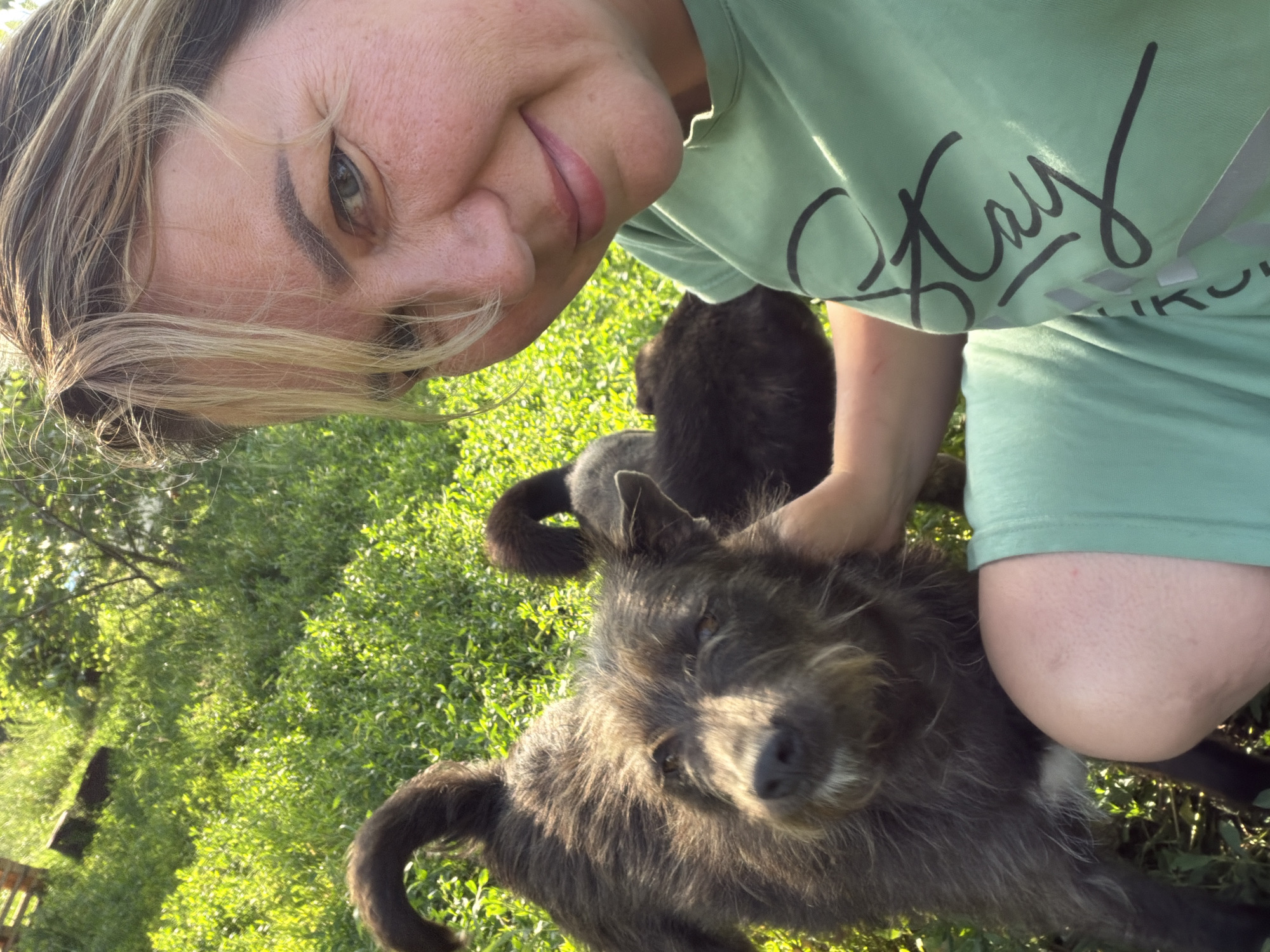 A woman, her face angled toward the camera in a selfie, smiles. She has blonde hair and is wearing a light green t-shirt with black lettering that appears to say "Stay." She is outdoors in a grassy area with trees in the background, suggesting a park or backyard. Sunlight illuminates the scene, indicating it's daytime, likely late afternoon due to the warm, golden light.
Two dogs are visible in the image. One is a dark-colored dog with scruffy fur, positioned in the foreground, looking towards the camera. Its tail is curled. Another darker, possibly black dog is partially visible behind the first dog. The woman's hand is resting on the head of the scruffy dog, suggesting interaction and affection between them. The overall mood is relaxed and happy, with the woman appearing to enjoy the company of her pets in an outdoor setting. The grassy area and foliage suggest a rural or suburban location.