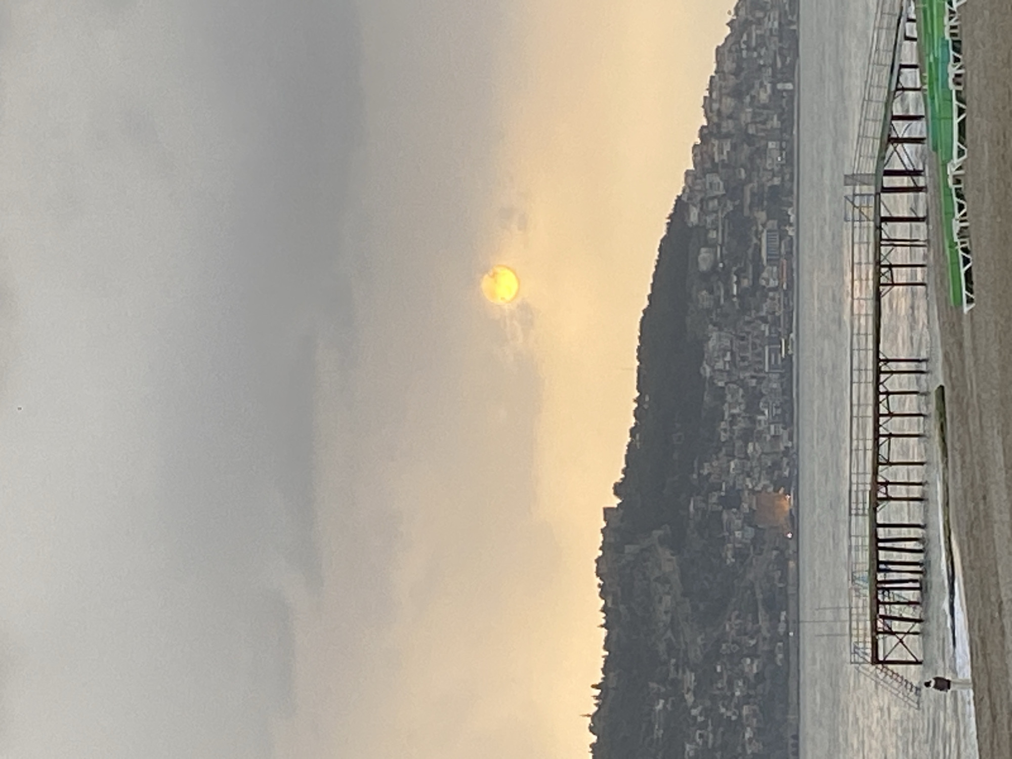 The image depicts a coastline at dusk. A pale, yellowish sun shines through a hazy sky. A large hill densely populated with buildings rises from the water's edge. A pier or structure extends into the water, with metal and wooden railings visible. A figure stands on the structure by the water. The location appears to be Alanya, Turkey.