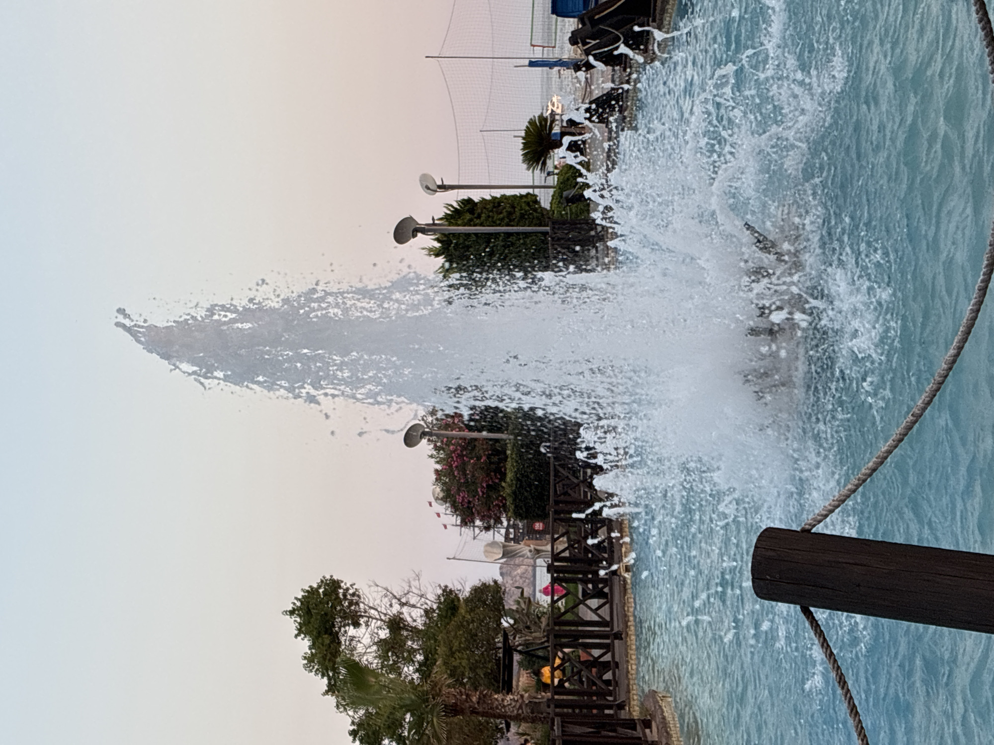 The image shows a water feature with a tall, powerful spray of water erupting from the center, creating a dynamic splash. The water is a bright, clear turquoise, suggesting it might be a swimming pool or a decorative fountain. In the background, a wooden railing lines the edge of the water, with a thick rope attached to a wooden post. Beyond the railing, there's a lush green landscape with trees and some flowering bushes. Several lampposts are visible, indicating that this area is lit, possibly for evening use. Further back, glimpses of structures and what appears to be a netting, perhaps for a sports court, can be seen. The sky is a soft, gradient of light blue and pale pink, suggesting either early morning or late evening. The overall scene conveys a sense of a recreational or resort area, likely in a warm climate given the vegetation. There are no people prominently visible in the frame, and the focus is on the water and the surrounding environment. The location is identified as Alanya, Turkey.