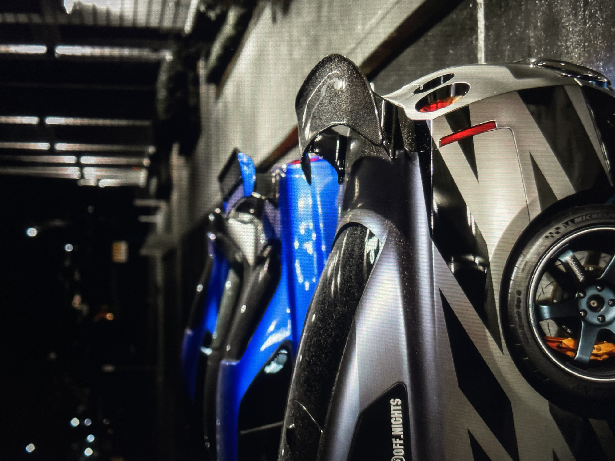 A close-up shot reveals a row of cars parked in what appears to be a garage. The cars are of various colors, including blue, gray, and black. The nearest car is gray with black and white diagonal stripes. The brand "OFF_NIGHTS" is visible on the car. The wheel of the gray car features a dark blue hubcap and an orange brake caliper. The background features metallic roofing and is dark and out of focus. The location is labeled Unknown, Unknown.