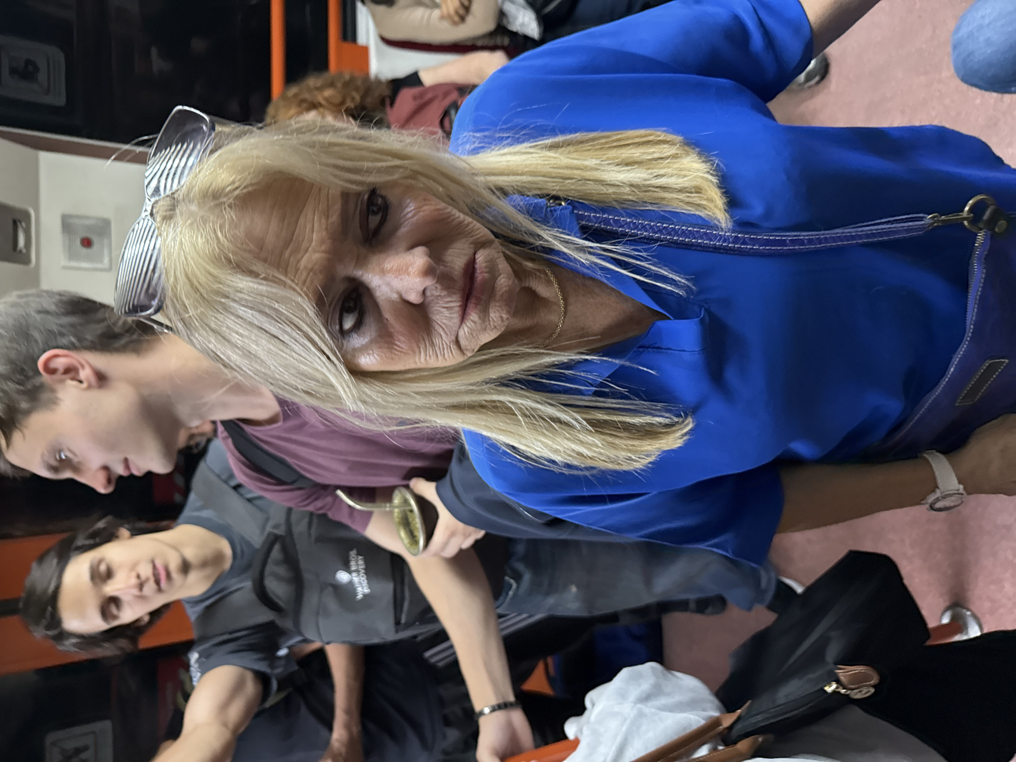 This indoor photograph, originally oriented sideways, depicts a crowded public space, likely within public transportation or a waiting area in Buenos Aires, Argentina.
The central figure is a blonde woman, appearing middle-aged, with visible wrinkles and a serious, somewhat displeased expression with pursed lips, looking directly at the camera. She wears a vibrant royal blue long-sleeved top and a delicate gold chain necklace. Stylish, striped sunglasses are pushed back onto her head, and a blue shoulder strap from a bag is visible. A white wristwatch is on her left wrist.
Behind and slightly to her left, a young man with dark hair, wearing a burgundy shirt, holds a traditional mate gourd with a metal bombilla, a strong cultural indicator for Argentina. Further back and to the left, another young man with dark hair wears a dark gray t-shirt featuring the text "WARNER BROS. DISCOVERY".
The setting appears to be an enclosed space with a reddish-pink floor. In the foreground, parts of a dark bag with a brown handle and a white cloth item are visible. In the background, elements of interior panelling and seating are discernible, along with a small red button or indicator light. The overall scene conveys an atmosphere of people traveling or waiting in a busy environment.