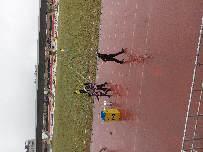 A sports field features runners on a red track next to a green field. A yellow and blue box sits near the track. A figure in black walks along the track. A stadium with visible seating is in the background, partially obscured by the overcast sky. A white fence section is visible on the right side of the image.