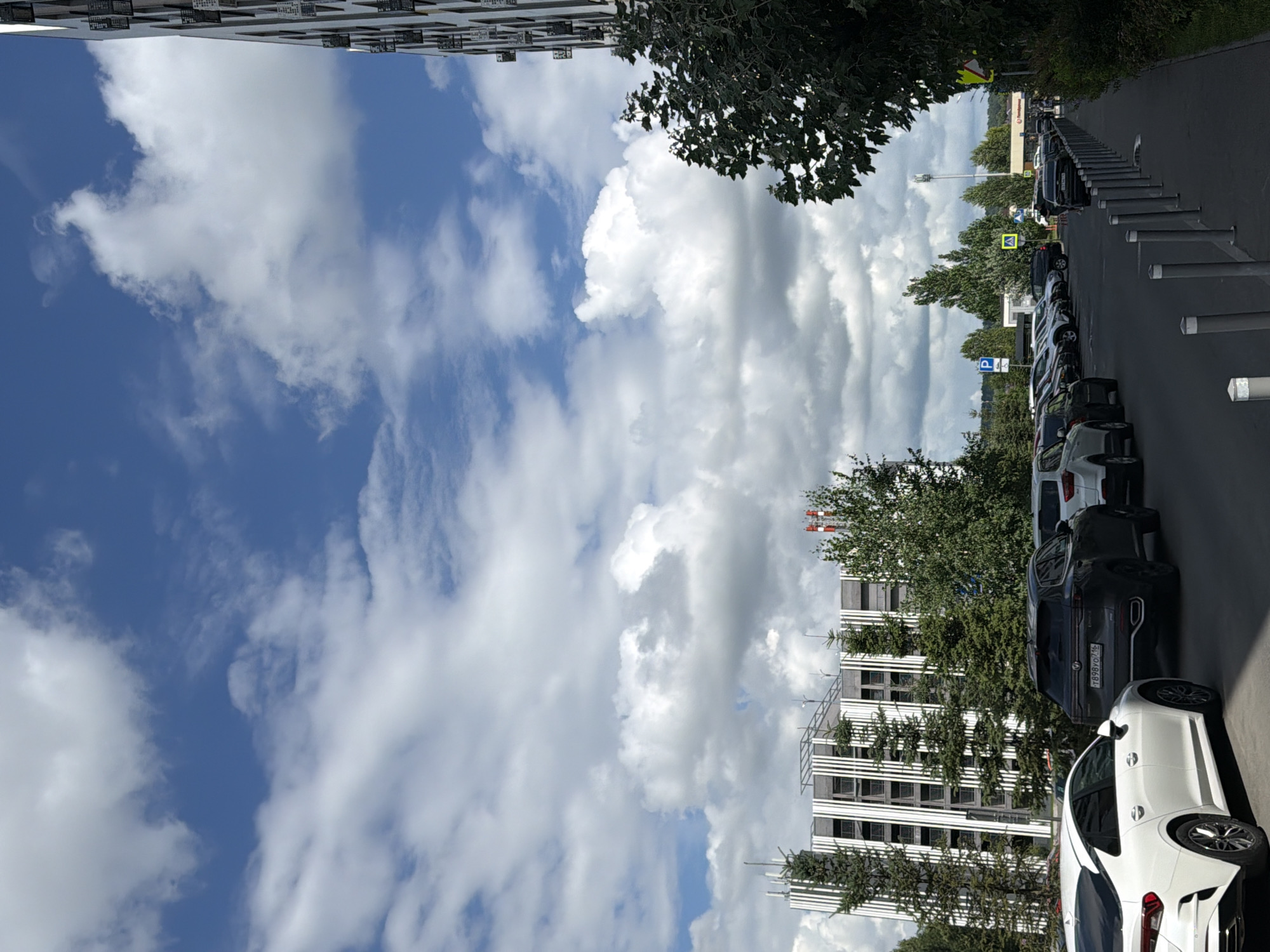 The image shows a parking lot with several cars lined up. In the background, there is a modern building with a white facade and vertical dark stripes. Trees, including some tall, slender evergreens, are planted in front of the building. The sky is partly cloudy with bright blue patches. To the right, there is a portion of another building with a dark facade and numerous small windows. A sidewalk with metal bollards runs along the right side of the parking lot. A yellow road sign with a black border is visible near the sidewalk. The license plate of the dark SUV in the foreground is partially visible and appears to be Russian. The scene is consistent with a commercial or residential area in Kazan, Russia.