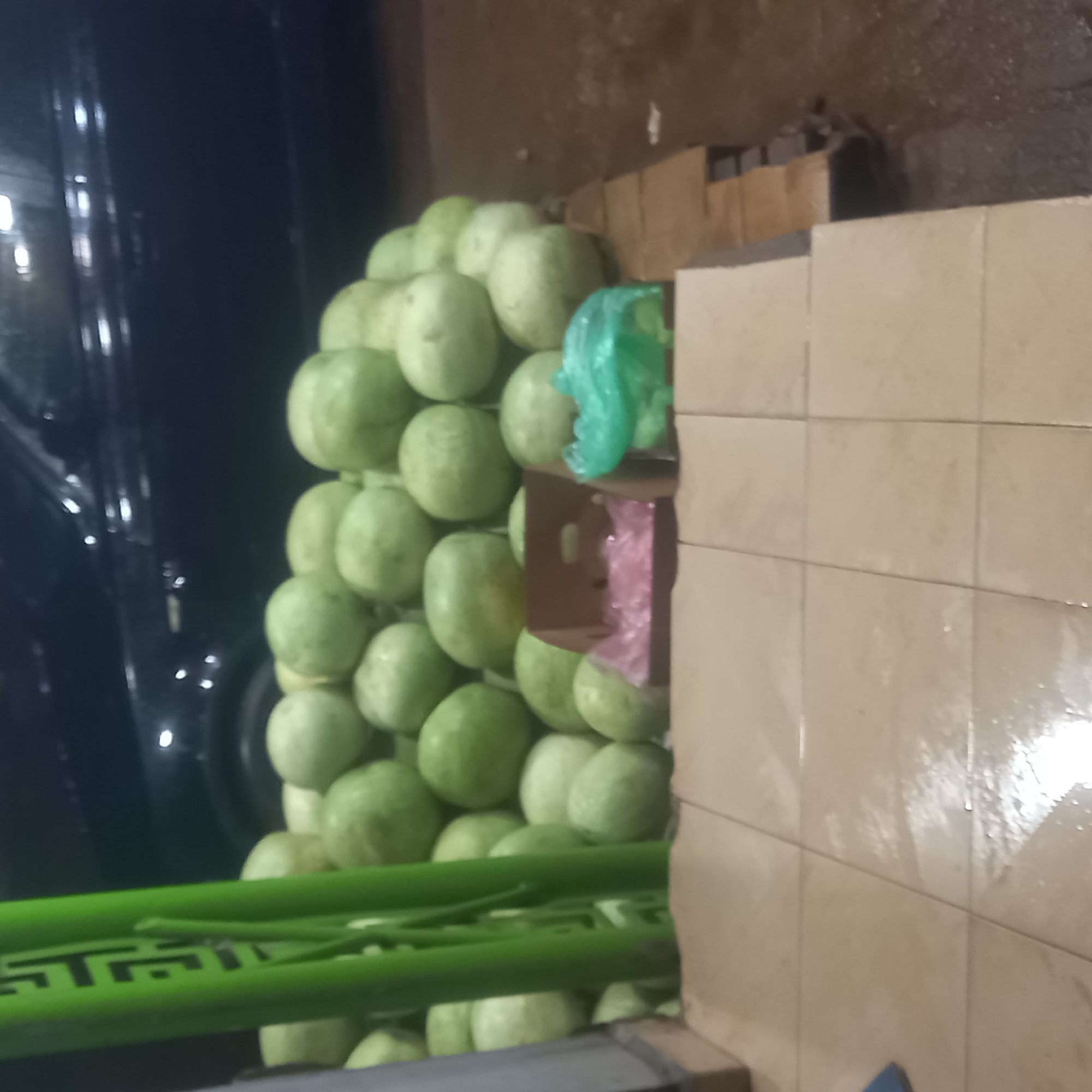A stack of light green, melon-like fruits is stacked next to a tiled wall. To the left of the stack is a green object. A dark vehicle is visible in the background. The location is possibly a marketplace or retail setting in Barnawa, Nigeria.
