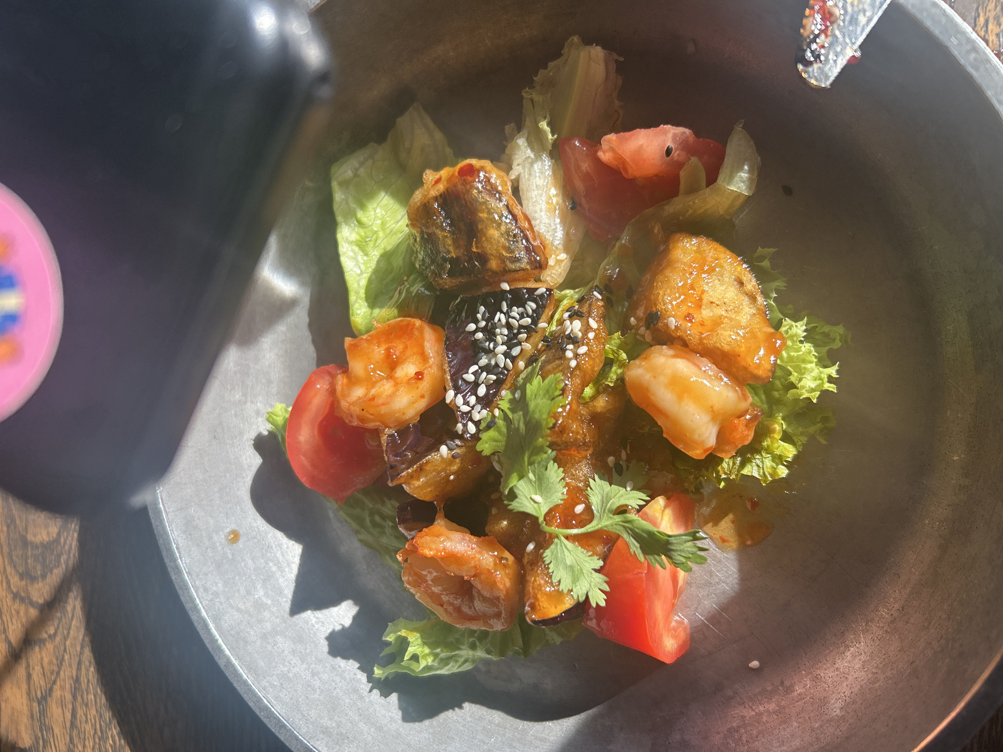 A close-up, overhead shot shows a dish of food served in a dark, metallic bowl. The food is composed of several shrimp, pieces of eggplant, and chopped tomatoes, all coated in a glossy, reddish sauce. The dish is garnished with cilantro and sprinkled with sesame seeds. The food is placed on a bed of lettuce. The bowl is set on a wooden table, with sunlight casting shadows. A black object, possibly a camera or phone, is visible at the top of the frame, partially obscuring the food and casting shadows. Another object, appearing to be a metal utensil with red remnants, is visible at the bottom right edge of the frame.