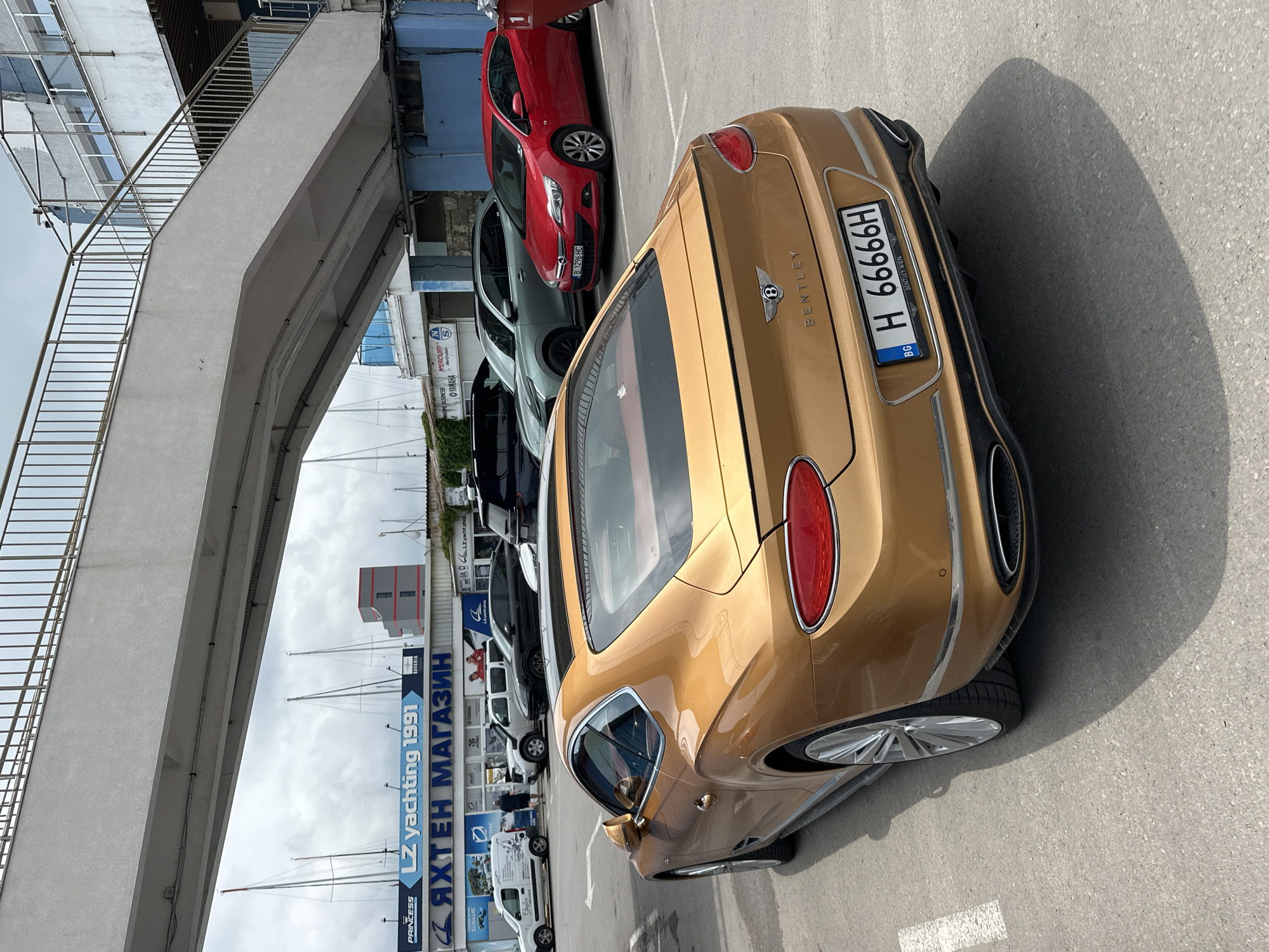 The image displays a sunny daytime scene in what appears to be a marina or port area in Varna, Bulgaria. In the foreground, a prominent gold Bentley Continental GT is parked, its rear facing the viewer. The car sports a Bulgarian license plate "H 66666H" with "BG" clearly visible, and "BENTLEY WIEN" is inscribed below the plate.
Behind the luxury vehicle, other cars are parked, including a red hatchback with a Bulgarian "B 1296 HC" license plate (indicating Sofia registration), a grey SUV or sedan, and several dark vans. To the left, a substantial concrete pedestrian overpass with metal railings extends upwards, suggesting a multi-level structure or passage over a road.
The background reveals the masts of numerous sailboats, confirming the waterfront setting. Buildings with modern blue glass facades and other structures are visible beyond the overpass. Several large banners and signs in both Cyrillic and Latin script are present, advertising yachting and marine-related businesses. Notable text includes "LZ yachting 1991" and "ЯХТЕН МАГАЗИН" (YACHT SHOP). Other visible brand names include PRINCESS, BAVARIA, SUBLUE, OYARANA, MERCURY, and YAMAHA, further reinforcing the nautical theme.
Bright sunlight illuminates the scene, casting distinct shadows on the asphalt parking lot. A few indistinct human figures can be seen near the buildings in the background, but no specific activity is occurring beyond the static arrangement of vehicles and commercial spaces. The scene conveys the atmosphere of a bustling coastal commercial hub.