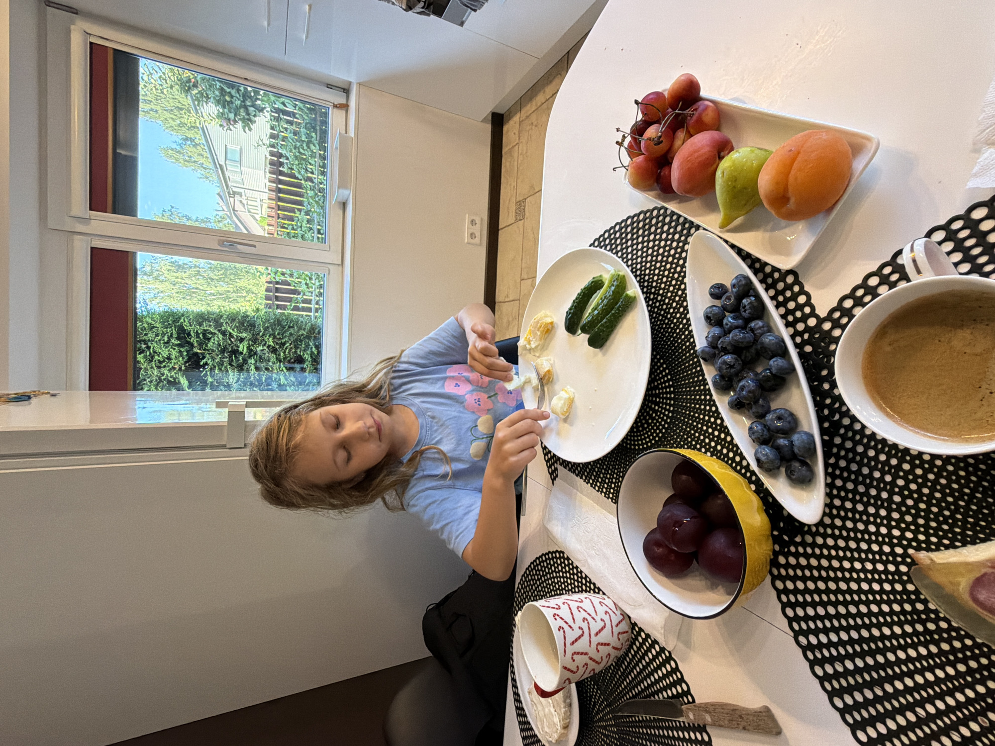 A young girl with blonde hair is sitting at a table in a modern kitchen, preparing her breakfast. She is wearing a blue t-shirt with a floral print and is focused on a plate of food. The table is set with various breakfast items, including a slice of bread with butter, a mug of coffee, a bowl of plums, a platter of blueberries, and a white rectangular plate containing sliced cucumbers, cooked egg pieces, and grapes, cherries, a peach, and an apricot. The girl is using a spoon to move pieces of egg on her plate. Behind her, a large window offers a view of a suburban neighborhood with houses, trees, and a blue sky. The kitchen appears clean and well-lit. The presence of a coffee maker and a small white container on the windowsill suggests this is a typical breakfast setting.