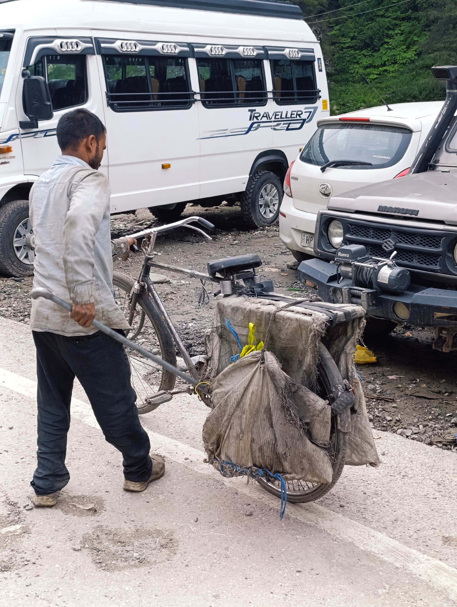 In Manali, India, a man is seen pushing a bicycle laden with a large, roughly wrapped bundle. The bundle appears to be covered by a heavy, canvas-like material, tied down with ropes and some yellow straps visible. The bicycle itself is old and utilitarian. The man is wearing a light-colored, long-sleeved shirt that appears dirty and possibly oil-stained, dark pants, and worn-out shoes. He is holding a metal pole that is extended and hooked onto the bicycle's frame, aiding him in pushing it.
The scene is set on a paved road with visible patches of dirt and gravel, suggesting it might be a less maintained area or roadside. In the background, several vehicles are parked. To the left is a white passenger van, identified by the "TRAVELLER 7" decal. To the right of the van is a white hatchback car. Further to the right, partially visible, is a rugged, off-road vehicle, possibly a Jeep, with a prominent winch mounted on its front bumper. The backdrop features lush green foliage, indicating a natural, possibly mountainous, setting. The lighting suggests it is daytime.