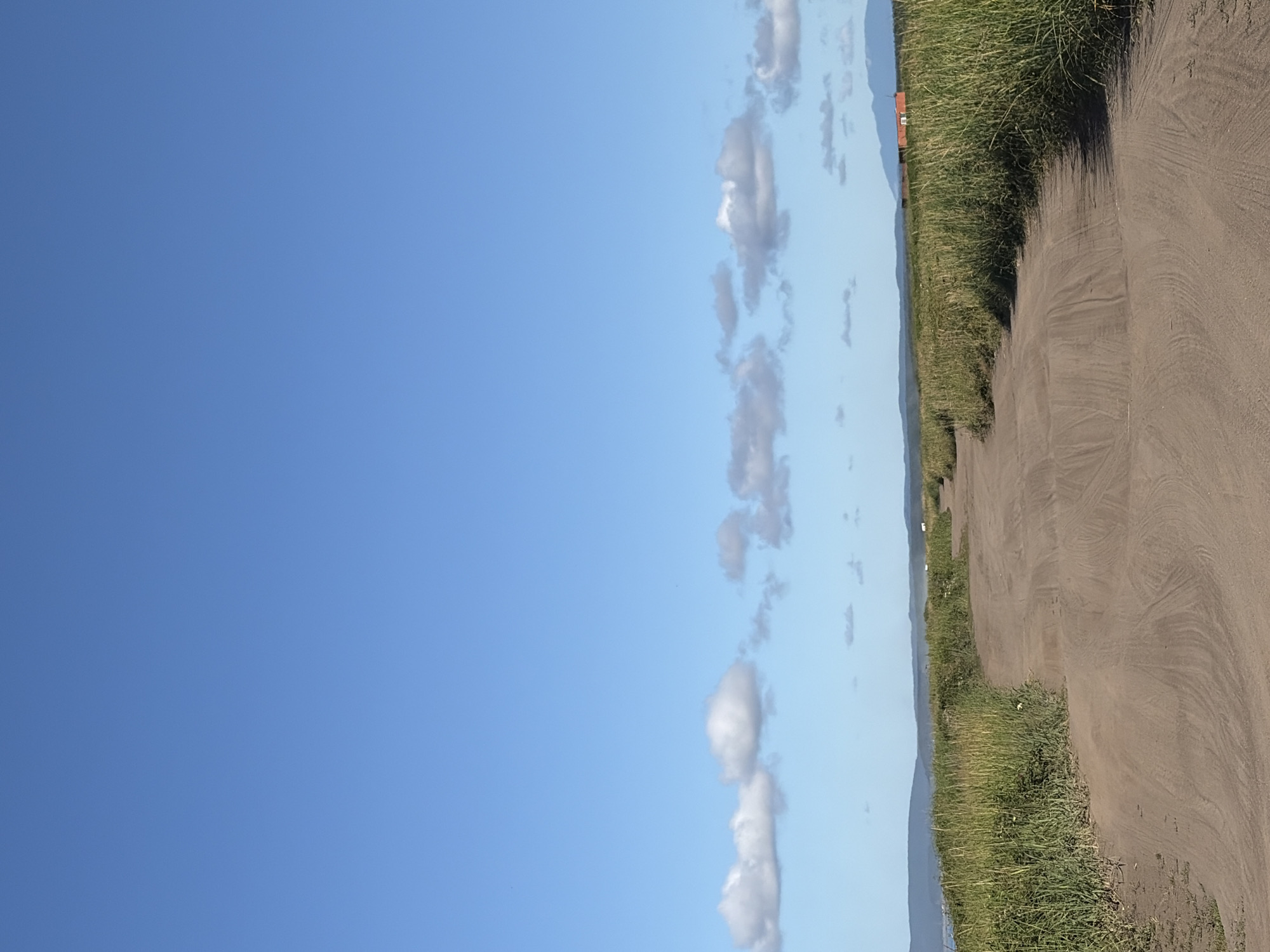 A dirt road curves through a field of tall, green grass under a clear blue sky. Scattered white clouds drift across the upper portion of the image. In the distance, rolling hills are visible, and a small, orange building with a dark roof sits on a distant rise. The road is packed dirt with visible tire tracks. The grass is thick and appears to be a mix of different species. The overall scene depicts a rural or natural landscape. The location is Ozerskoe, Russia.