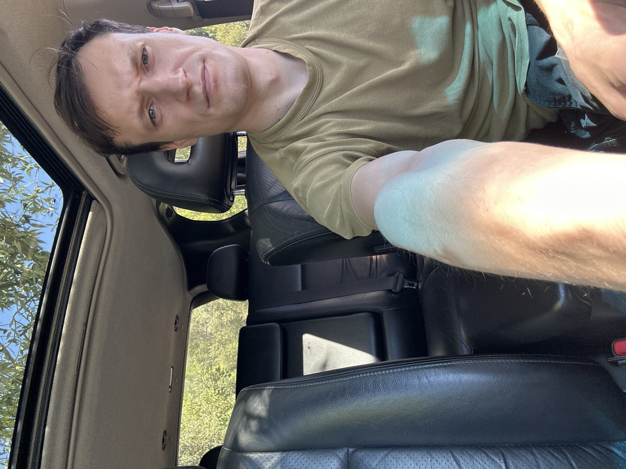 A man with short brown hair and a light complexion is sitting in the driver's seat of a car, looking directly at the camera with a neutral expression. He is wearing a tan t-shirt and dark shorts. The car's interior is visible, including black leather seats, a sunroof, and a portion of the dashboard. Sunlight is streaming in through the open sunroof, illuminating the interior and casting shadows. The car appears to be parked outdoors, with blurred green foliage and a blue sky visible through the windows. The image was taken in the city of Makiv, Ukraine.