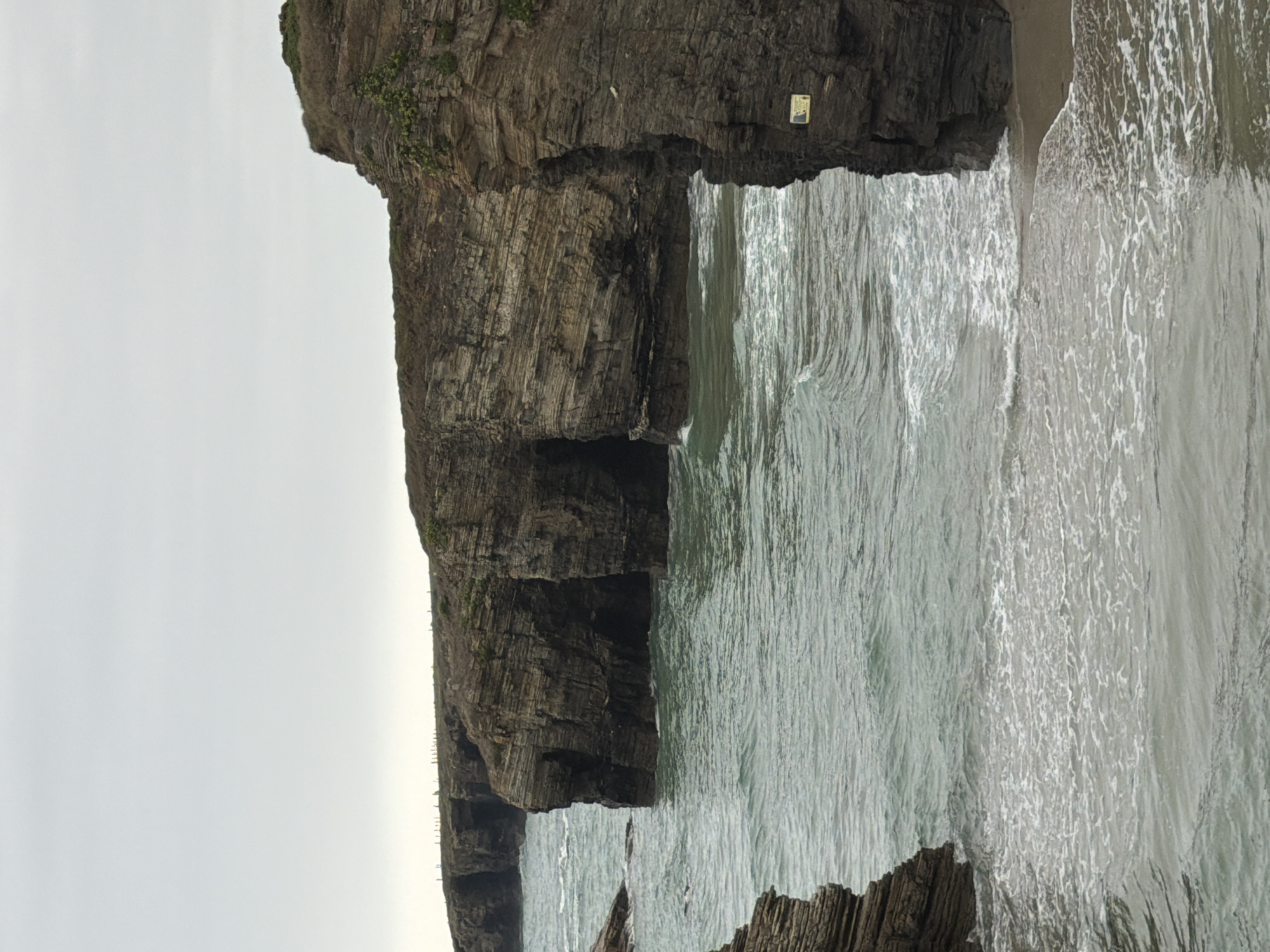 The image shows a view of the Cathedrals Beach in Ribadeo, Lugo, Spain, under an overcast sky. The foreground features gentle waves lapping onto a sandy shore. The dominant features are the natural rock formations and arches carved by the sea. These rock formations are layered and weathered, with vegetation growing on top in some areas. In the middle ground, the sea flows between the rock structures, creating a sense of depth and movement. The background shows a continuation of the rugged coastline, with more rock formations extending into the distance and a thin line of fence posts along the cliff edge. One of the rock faces in the right middle ground has a small, yellow sign attached to it, but the text is illegible. There are no people or animals visible in the scene. The overall mood is one of natural beauty and the power of the ocean.