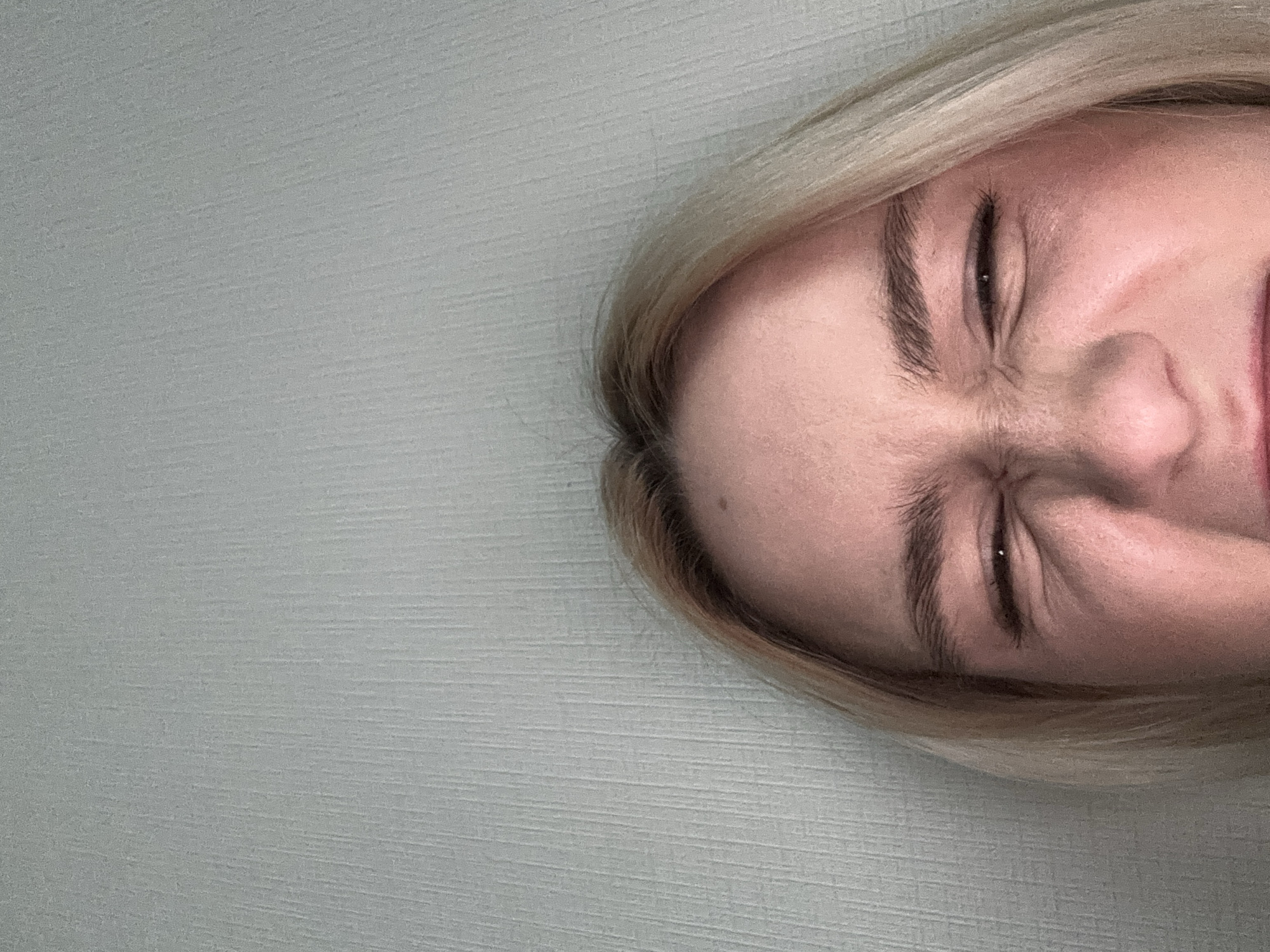The image shows a close-up of a person's face with eyes tightly shut, positioned against a plain wall. The person has fair skin and blonde hair. The wall appears to be a light neutral color, possibly with a textured surface. There is no information in the image to suggest a location in Vsevolozhsk, Russia, or any identifying features about a specific city. The image primarily captures a facial expression and provides limited contextual details.