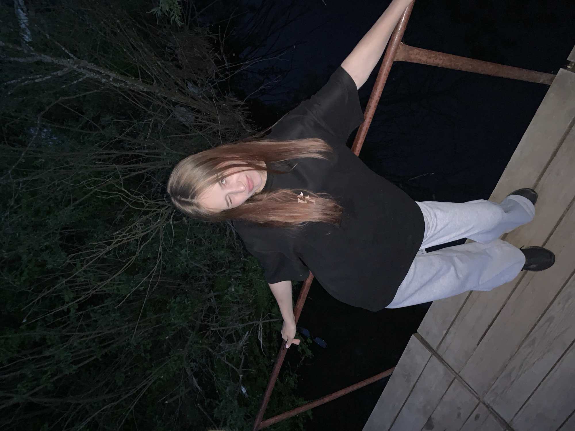 A young woman with long brown hair stands on a wooden platform in what appears to be Фоки, Russia, at night. She is holding onto a rusty metal railing. She is wearing a black shirt, light grey sweatpants, and dark shoes. She is also holding a pair of sunglasses in her left hand. The background consists of trees and a dark body of water. The scene is dimly lit.