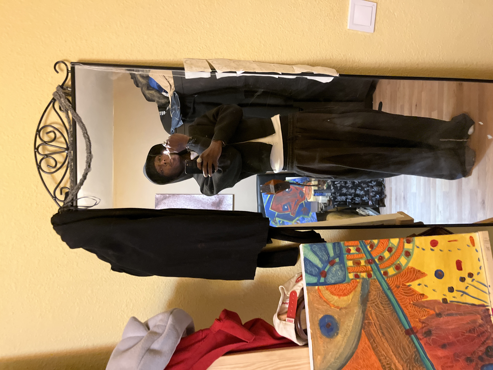 A reflection of a person in a mirror dominates the image. The person in the reflection is wearing dark clothing and appears to be taking a picture with a cell phone. The background reflected in the mirror includes a wooden floor and miscellaneous items. Adjacent to the mirror on the right side of the frame is a colorful painting. The wall behind the mirror is a muted yellow color. There is no information in the image that indicates the location is in Barcelona, Spain.