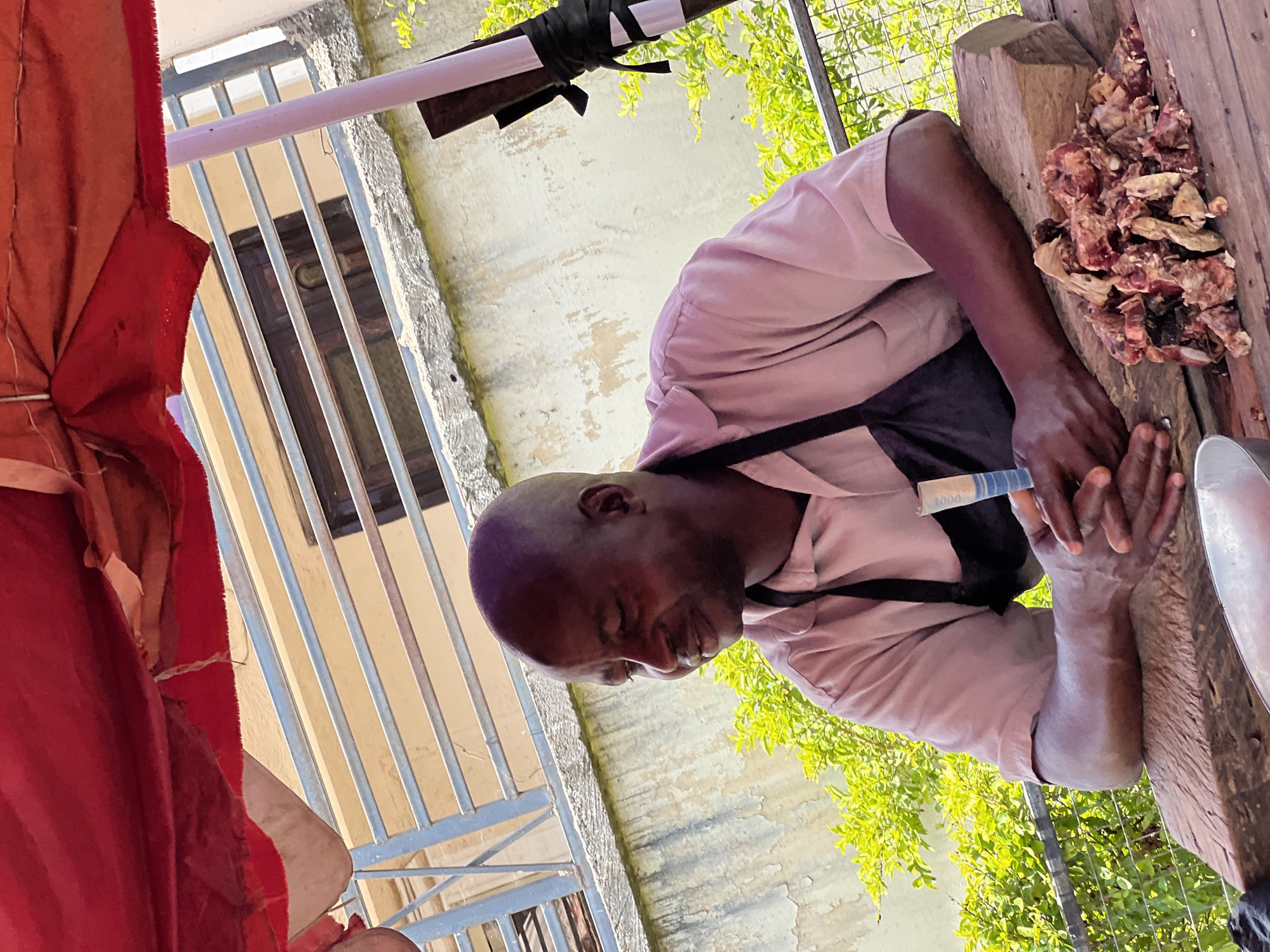 This image depicts a man actively preparing meat at an outdoor or semi-outdoor stall, likely in a market setting in Jos, Nigeria. The main subject is a dark-skinned man with a bald head, dressed in a light pink short-sleeved shirt and a dark apron. He is positioned over a large, well-worn wooden chopping block or table, where chunks of raw, bone-in meat are piled. His hands are engaged with the meat, suggesting he is cutting, chopping, or arranging it. A small metal bowl or pan is visible on the right side of the wooden surface.
The scene is illuminated by bright natural daylight, indicating it is daytime. To the left, a vibrant red fabric, possibly part of a canopy or tent, partially frames the image. In the background, there are metal railings, suggesting a fence or balcony. Beyond these railings, a light-colored wall with some weathered patches is visible, alongside lush green foliage from trees or bushes. A light-colored pole, secured by a black strap, runs horizontally in the upper part of the frame.
A notable detail is a partially visible banknote in the man's left hand, clearly showing the number "1000", which identifies it as a 1000 Nigerian Naira note, implying a transaction has taken place or is about to. The man appears focused and industrious, dedicated to his task of meat preparation. The overall impression is that of a bustling, functional market environment.