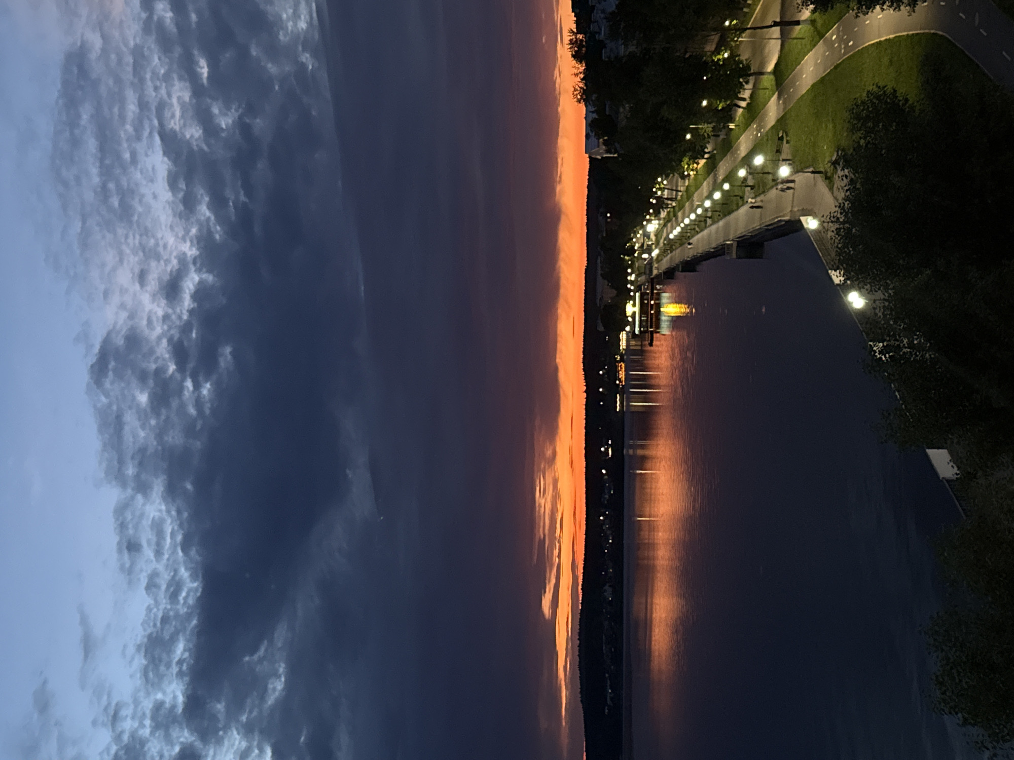 An aerial shot captures a twilight scene along the Votkinsk Reservoir embankment in Votkinsk, Russia. The sky is a deep blue, transitioning to a vibrant orange and red horizon as the sun sets. Dark, textured clouds dominate the upper portion of the sky. The calm water of the reservoir reflects the fiery colors of the sunset and the lights from the embankment.
Along the curved embankment, a paved walkway is illuminated by numerous evenly spaced streetlights. Green grass borders the walkway, and a separate paved path, possibly for cycling or jogging, curves alongside it. Trees line the embankment, their dark silhouettes contrasting with the illuminated walkway. A few indistinct figures can be seen walking along the promenade. Buildings and trees are visible on the distant shoreline, with scattered lights indicating a populated area. One building on the embankment features glowing yellow lights. The overall scene depicts a peaceful evening in a city park setting.