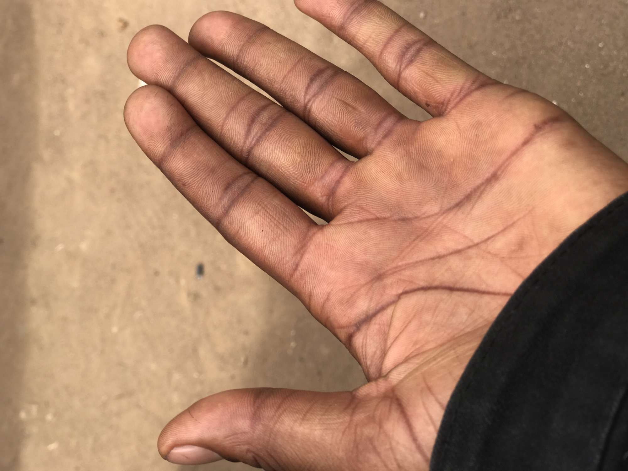 The image displays a close-up view of the palm of a person's left hand. The hand is positioned with fingers pointing upwards, and a black fabric sleeve is visible covering the wrist area on the right side of the frame. The skin tone is dark, and the surface of the palm and fingers appears to be coated with a thin layer of dust or dirt, giving it a somewhat dull, brownish-greyish complexion. The natural creases and lines on the palm and finger joints are highly pronounced, appearing darker, almost purplish-brown, indicating they may be stained from exposure to foreign substances. Detailed fingerprint patterns are visible on the tips of the fingers. The background is out of focus, showing a light brown or beige textured surface, possibly a floor or ground. No specific activity is taking place, but the condition of the hand suggests manual labor or interaction with a dusty environment. No text is visible in the image.