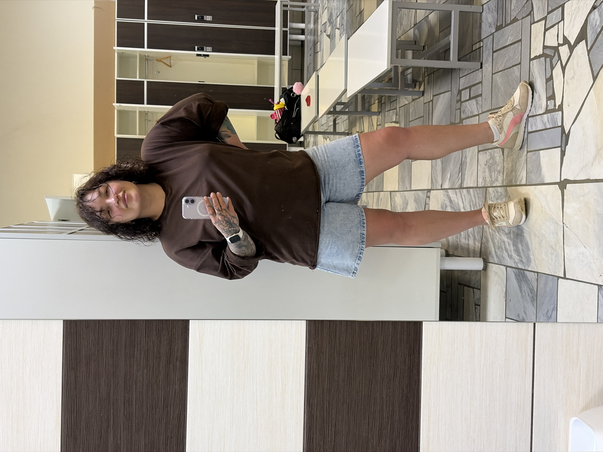 A woman is standing in a locker room and taking a selfie. She is wearing a brown sweatshirt and light blue denim shorts. She has dark, curly hair and tattoos on her arms. In the background, there are rows of lockers and benches. The floor is tiled with a pattern of grey, white, and beige tiles. The lighting is bright and even. This scene is likely in Chaikovsky, Russia, given the context of the user's request.
