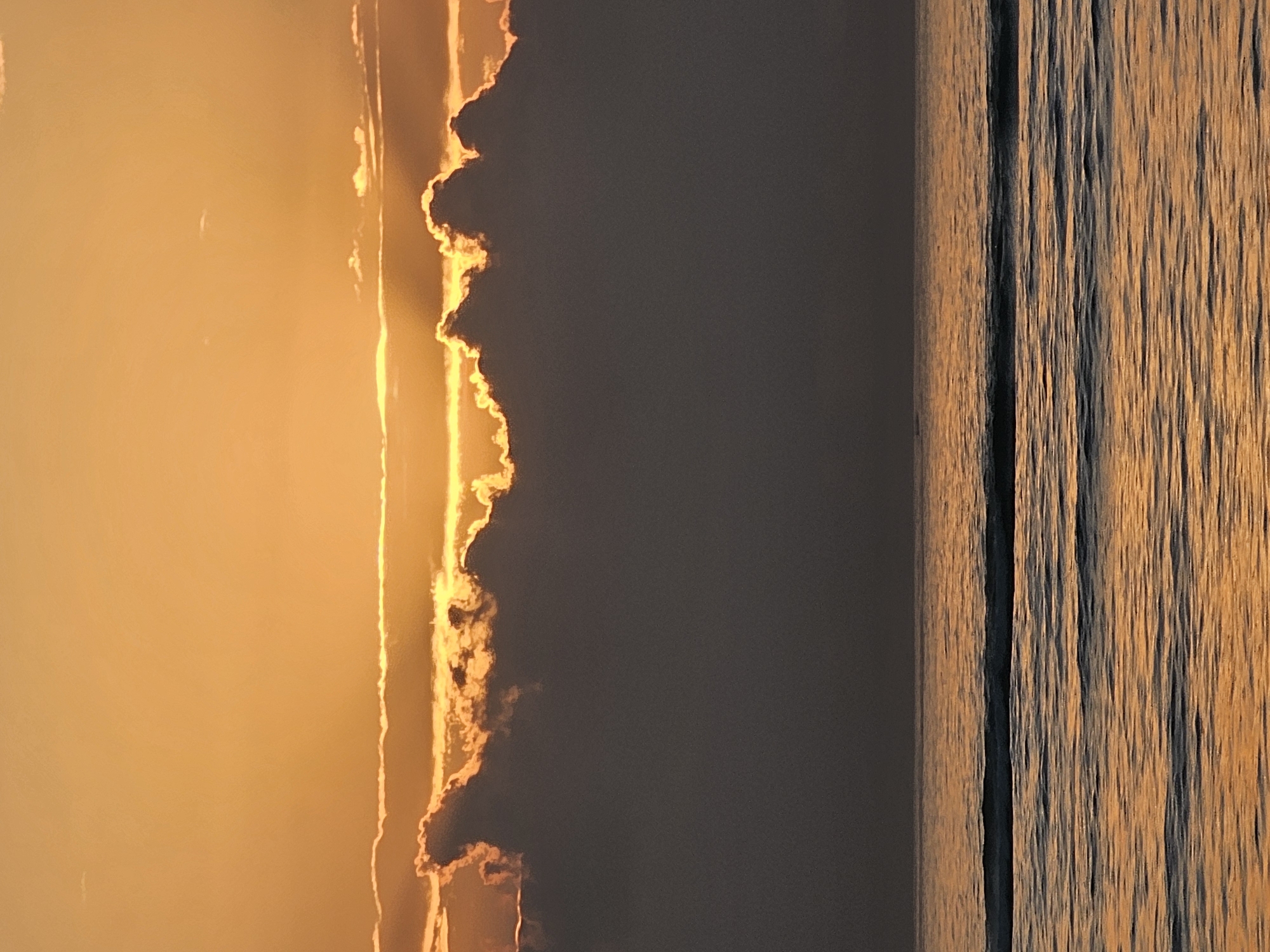 The image depicts a sunset over the ocean, likely at Seminyak Beach in Denpasar, Indonesia. The sky is filled with vibrant orange and yellow hues, indicating the sun is setting near the horizon. Dark, silhouetted clouds are positioned in the middle ground, partially obscuring the glowing sunlight that peeks through their edges. Below the clouds, the ocean is visible, with gentle waves reflecting the warm colors of the sunset, creating a shimmering, golden surface. The horizon line is clearly defined where the sea meets the sky. No people or man-made structures are visible in the frame, focusing solely on the natural beauty of the seascape at dusk.