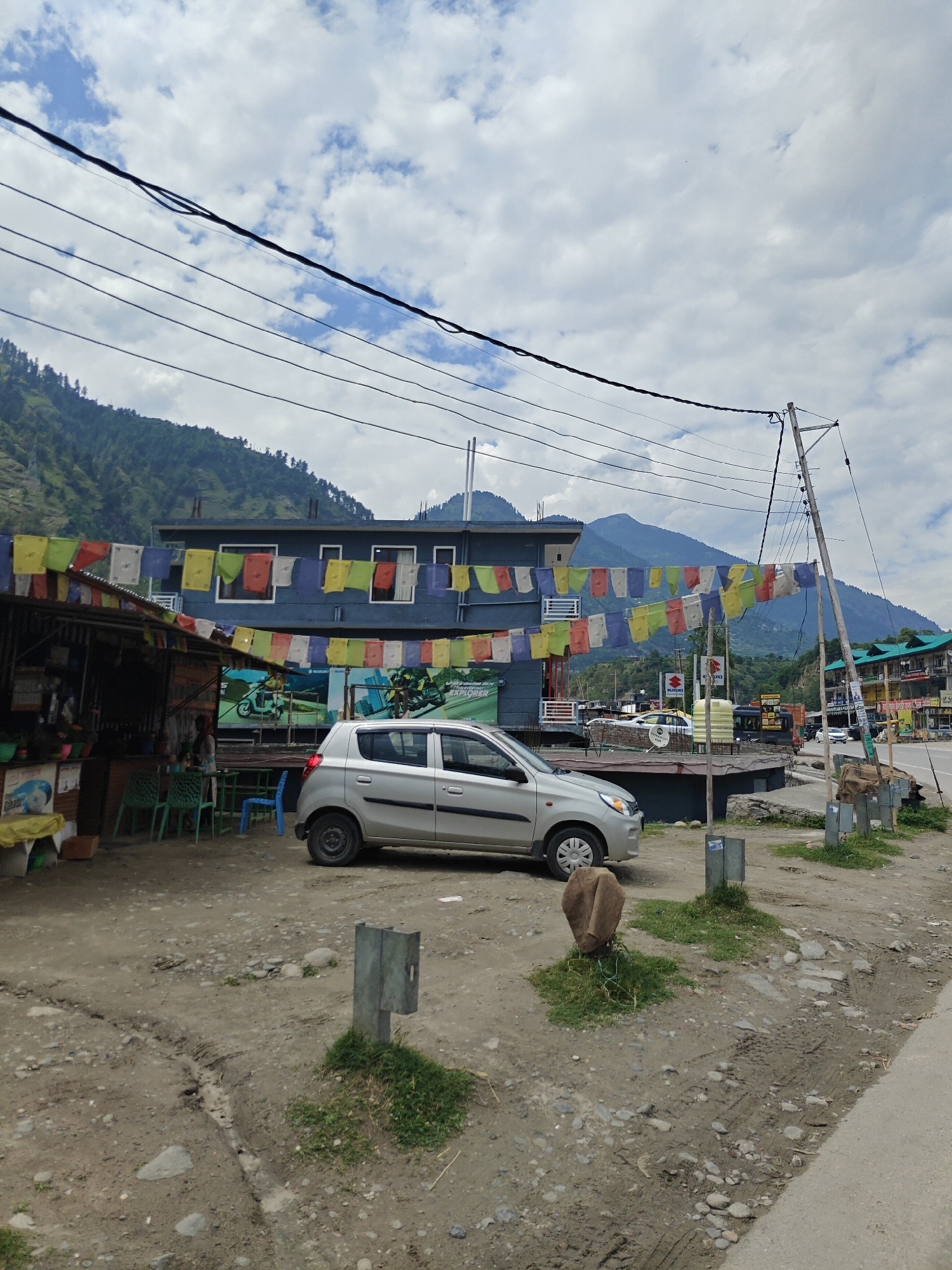 This image captures a daytime scene along a roadside in Patlikuhal, India, set against a backdrop of lush green mountains under a cloudy sky.
In the foreground, the ground is uneven, composed of dirt and gravel with small patches of grass. A light silver hatchback car, possibly a Maruti Suzuki Alto, is parked facing right. To its left, partially obscured, is a small, rustic roadside stall or shop with an awning, where a person is visible behind a counter. Several green plastic chairs and one blue chair are arranged outside the stall, suggesting a seating area. Above this stall and stretching across the scene, colorful Tibetan prayer flags are strung, adding a vibrant cultural element.
Behind the car and prayer flags, a modern-looking building with a dark facade and white-framed windows is visible. A large banner or poster on this building features motorcycle imagery and the visible text "SUZUKI." Further to the right, a tall utility pole stands beside the road, which curves into the distance. More buildings, some with green roofs, and other vehicles can be seen along the road in the midground. Another sign with "SUZUKI" and possibly "BAJAJ" is visible on a building to the far right.
The background is dominated by towering, tree-covered mountains, indicating a mountainous or hilly region. The sky is largely overcast with scattered white clouds, suggesting a mild or pleasant day. No specific activities are prominently displayed, but the setup suggests a regular roadside stop or commercial area. The overall impression is that of a bustling yet scenic location in a Himalayan foothills town.