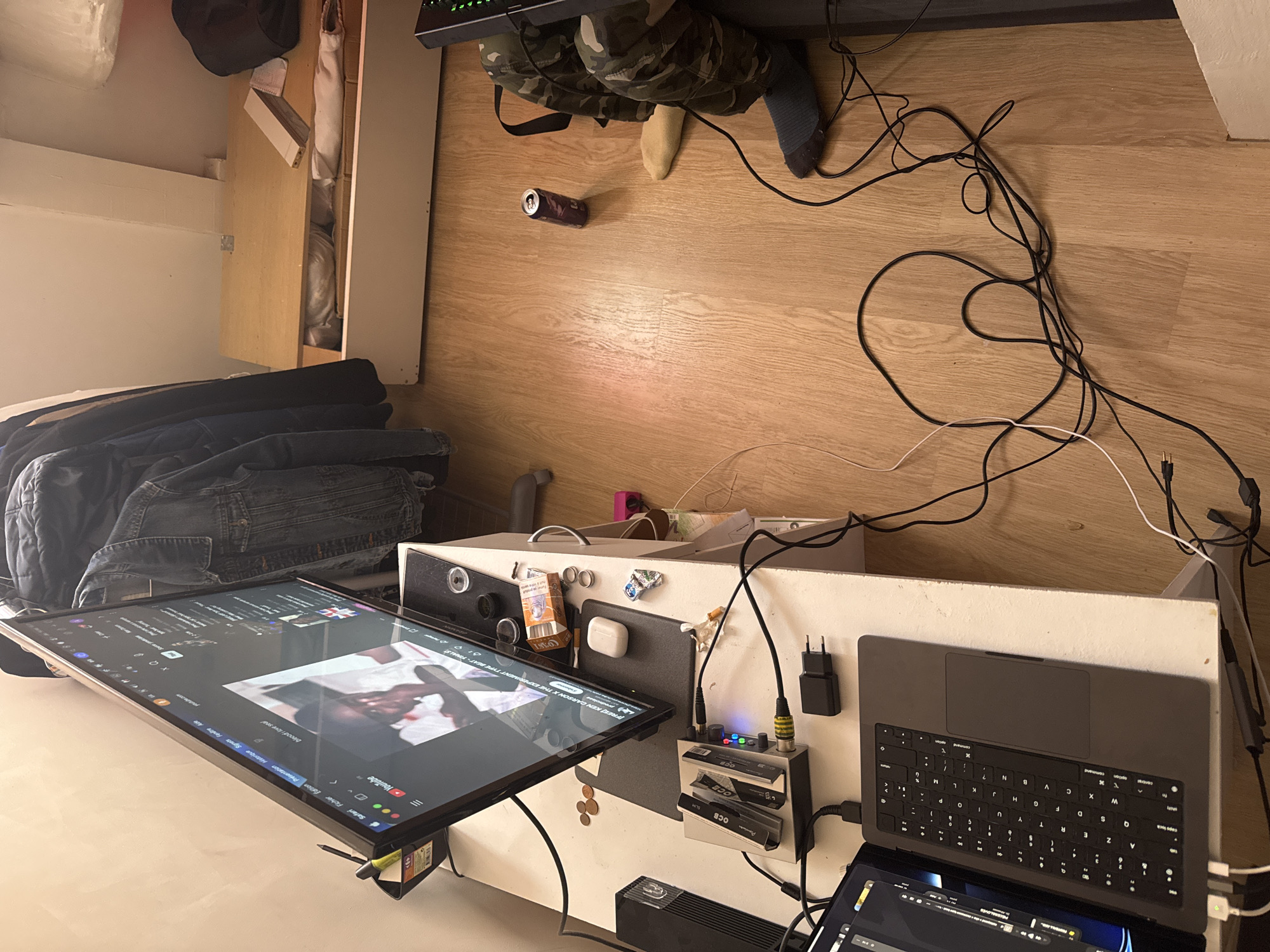The image shows an untidy desk setup, likely in a domestic setting. The desk is white and covered in electronic devices, including a large monitor displaying a YouTube video, a laptop, an audio interface (OCB slim audio interface), and various cables. Several small objects, such as coins, a pack of cigarettes, and Apple AirPods, are scattered around the desk's surface.
On top of the desk, clothes are piled. On the floor, there are additional items, including an empty can and a person wearing camouflage trousers and mismatched socks. A significant amount of tangled cables is strewn across the floor.
The environment suggests an indoor location, possibly a home office or bedroom. The background includes a wooden floor and a glimpse of a wardrobe or storage unit.
There's no indication this is in Paris, France.