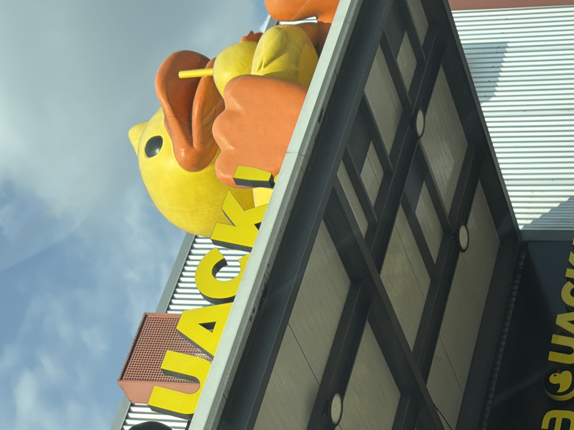 The image shows a large, stylized yellow duck figure with an open orange beak holding a yellow popsicle stick, mounted on a building with the name "QUACK". The building has a modern architectural design with a dark-colored crossbeam pattern. The sky with clouds is visible behind the duck. The photograph was taken from a low angle. This is the famous “Pollos Quack” restaurant in San Pedro Cholula, Mexico.