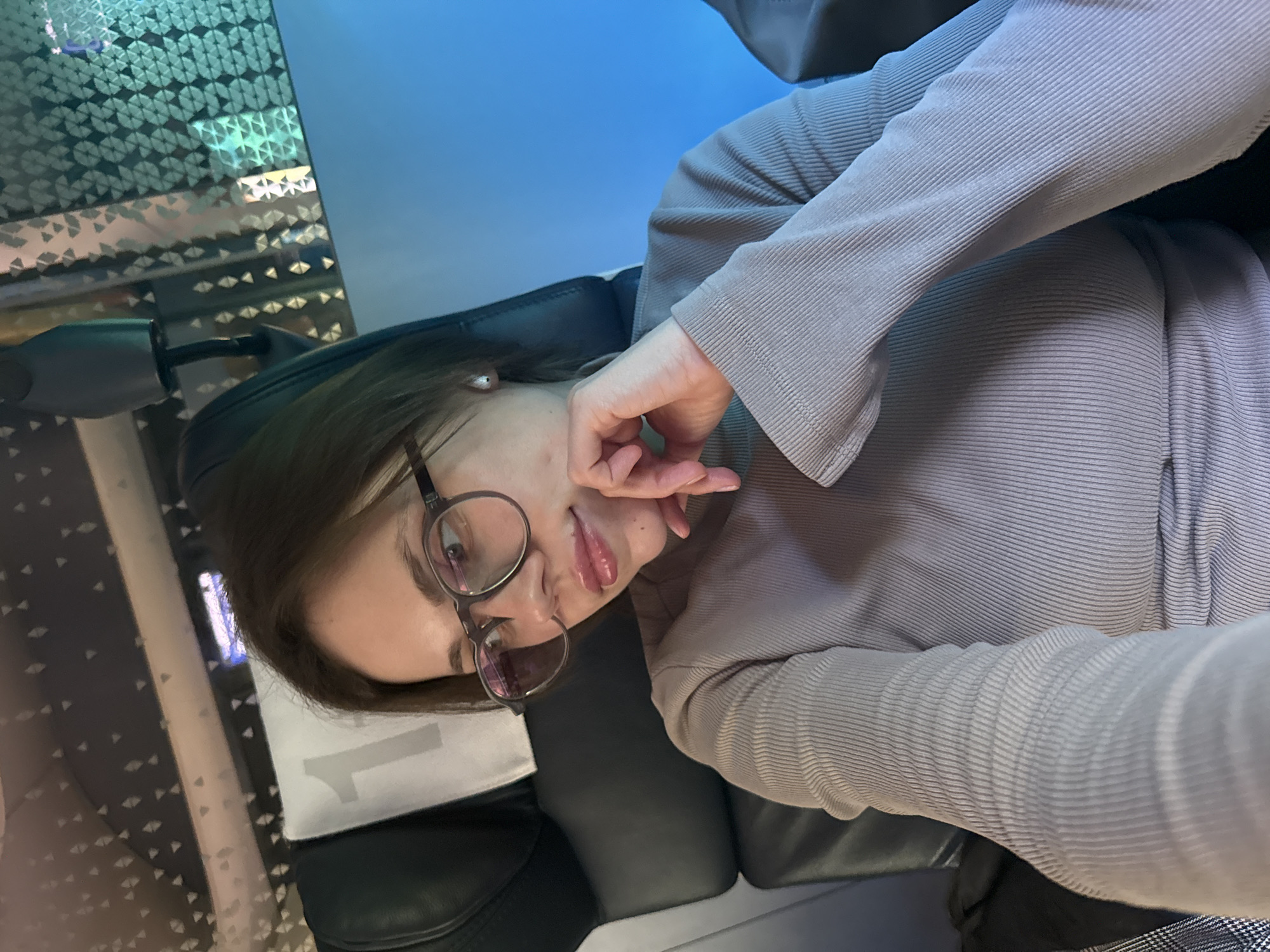 The image shows a person with brown hair and glasses, wearing a light gray long-sleeved shirt, sitting in a black gaming chair. The background is blurred and features a combination of blue light and a geometric pattern resembling a wall. The location is described as Unknown, Unknown. The person's hand is near their face in a contemplative gesture. The camera angle is slightly tilted.