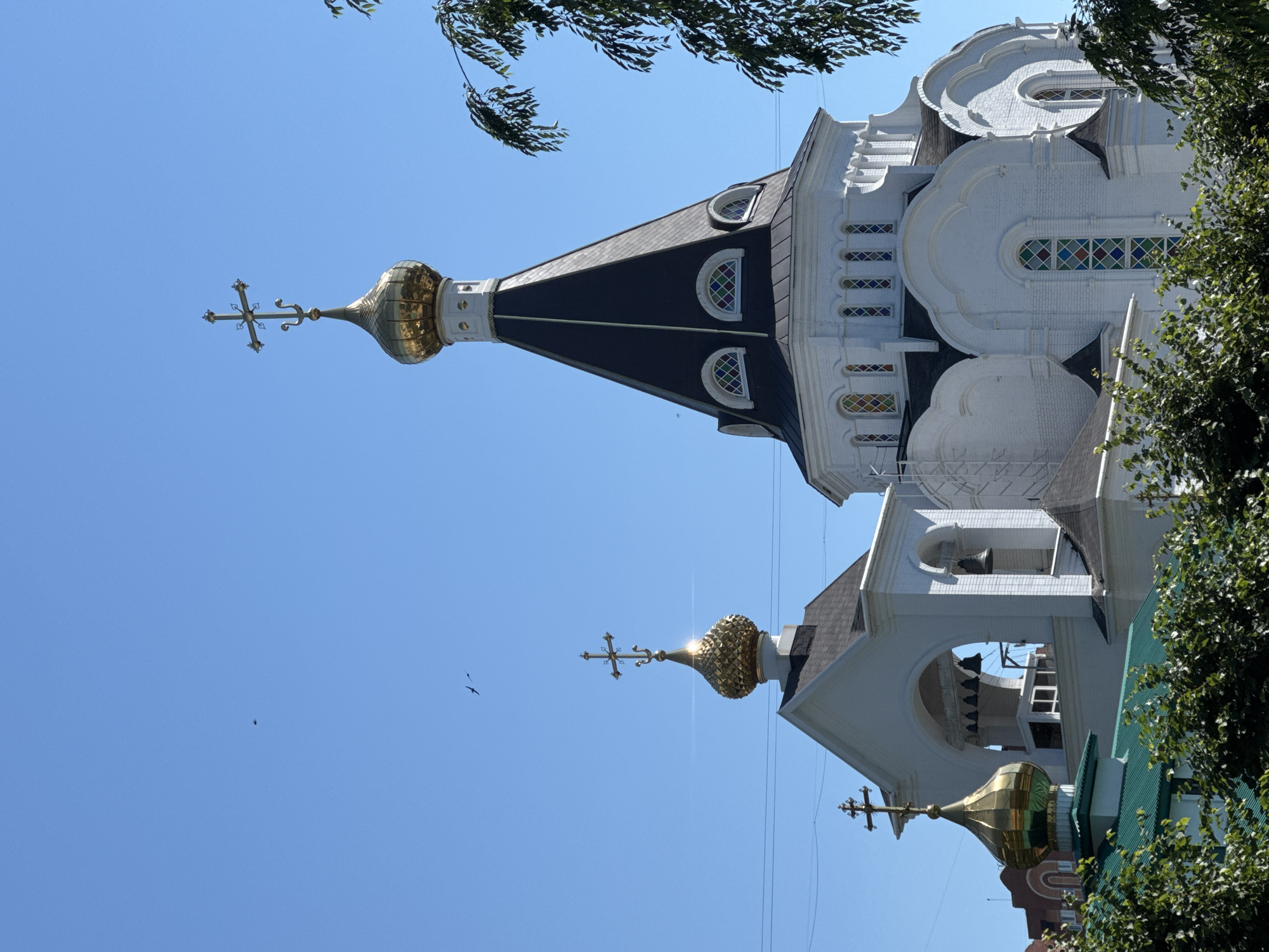 A low-angle, full-shot photograph captures the exterior of an Orthodox church against a clear blue sky. The white brick building features multiple golden domes topped with crosses, and a tall, dark, conical roof with arched windows. The structure exhibits architectural details such as arched doorways, decorative trim, and stained-glass windows. The foreground is partially obscured by the lush green foliage of trees and bushes. Two birds are visible in flight in the upper left quadrant of the sky.