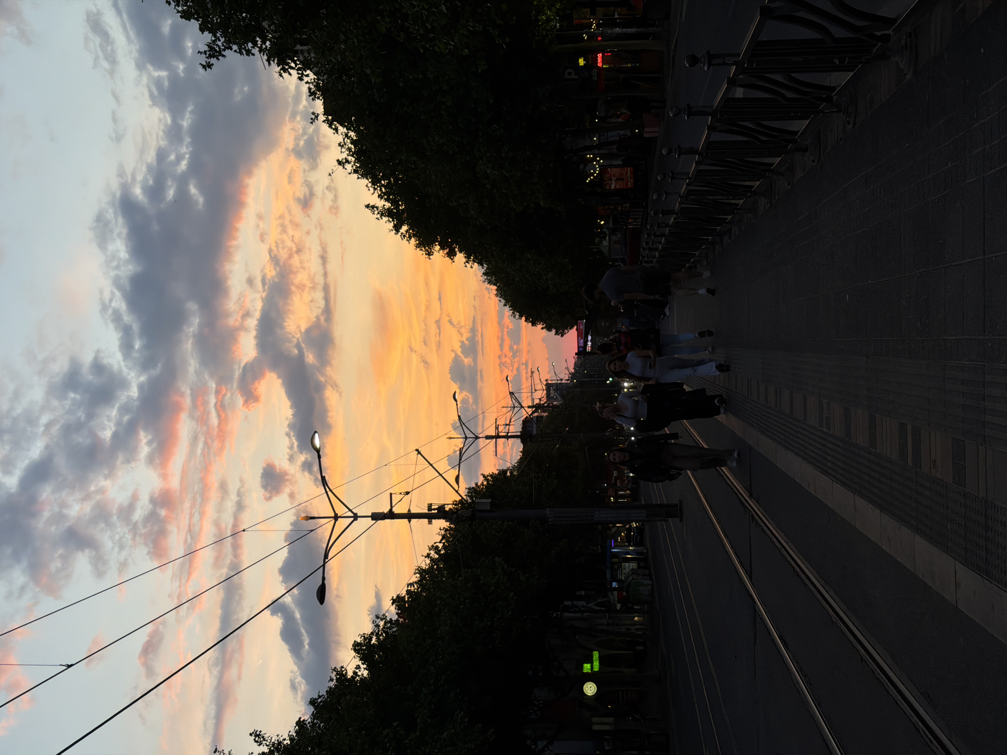 The image depicts a street scene in Belgrade, Serbia at dusk. The sky features a colorful sunset with orange and gray clouds. A tram track runs along the ground, with pedestrians walking on the sidewalk alongside it. Overhead power lines and streetlights are visible. Trees line the street. A metal railing is seen along the side. People are present, some walking and others standing. The environment appears to be urban, with buildings and storefronts faintly discernible.