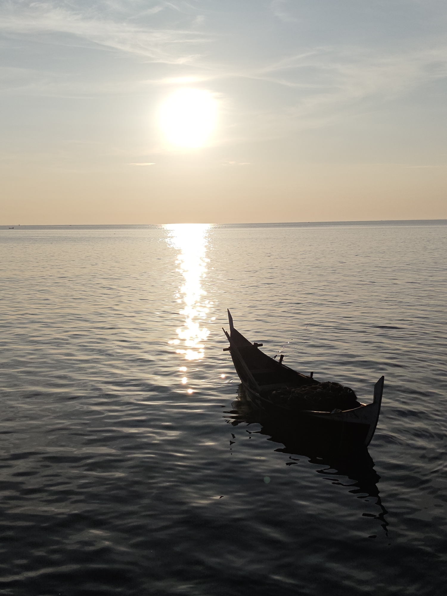 A silhouette of a traditional wooden fishing boat is captured on the calm waters of Lhokseumawe, Indonesia, during sunset. The sun, a bright orb, is positioned high in the sky, casting a shimmering reflection across the sea. The sky transitions from a pale blue at the top to a warm yellow and orange hue near the horizon, indicating the approaching evening. The water is dark and reflects the warm tones of the sky and the bright sun. The boat, dark and elongated, appears to be empty and is slightly tilted, suggesting it is at rest. It contains a pile of what appears to be fishing nets or similar equipment in its center. The overall scene is tranquil, with the gentle ripples on the water adding a sense of peaceful movement.