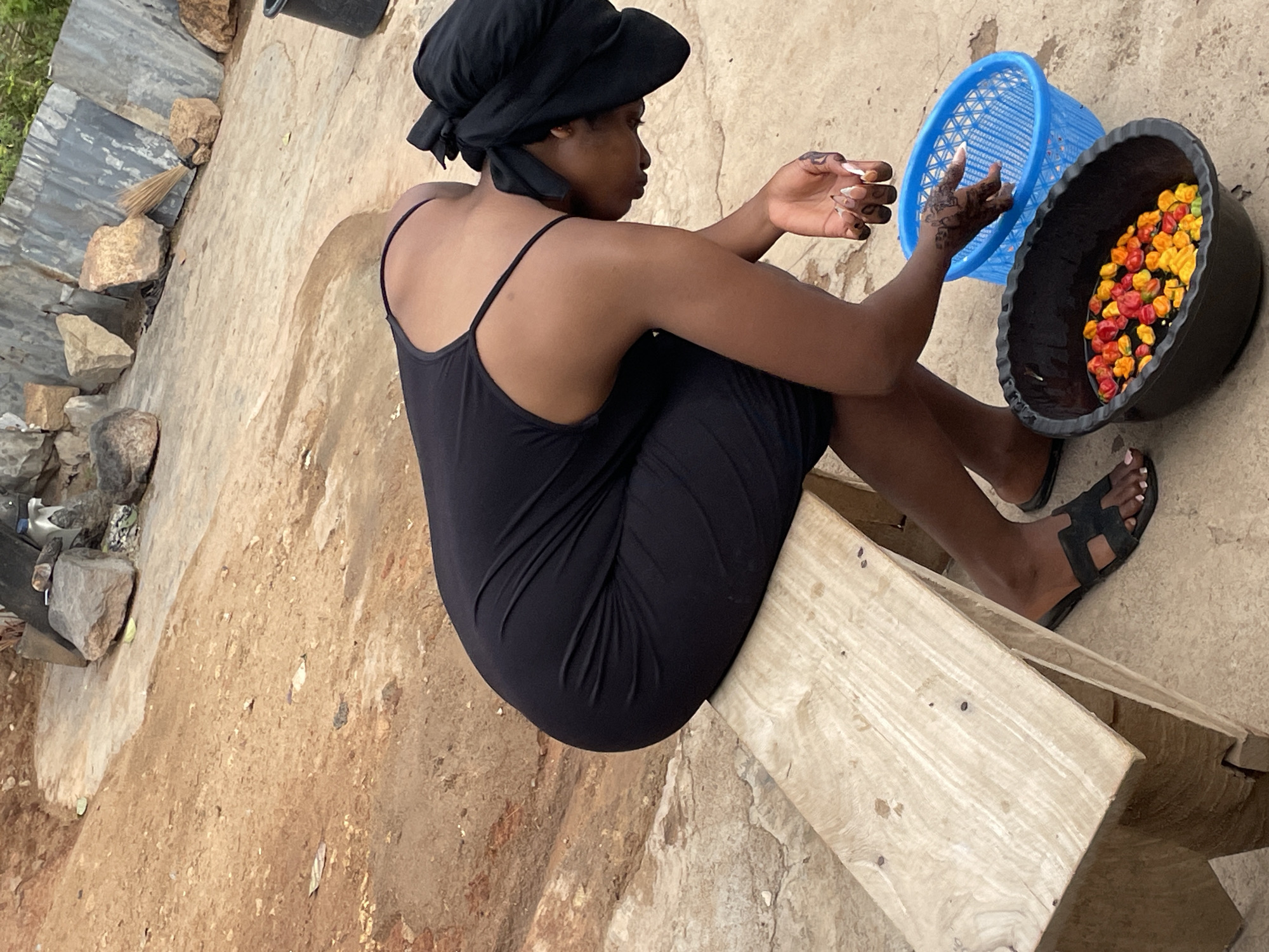 A woman wearing a black head covering and a black dress is sitting on a wooden structure outside a building in Jos, Nigeria. She is sitting next to a black bowl filled with colorful peppers and a blue basket. Her hands are raised in front of her, appearing to sort or handle the peppers. She is wearing black sandals. The ground is a mix of dirt and concrete. The building has corrugated metal siding.