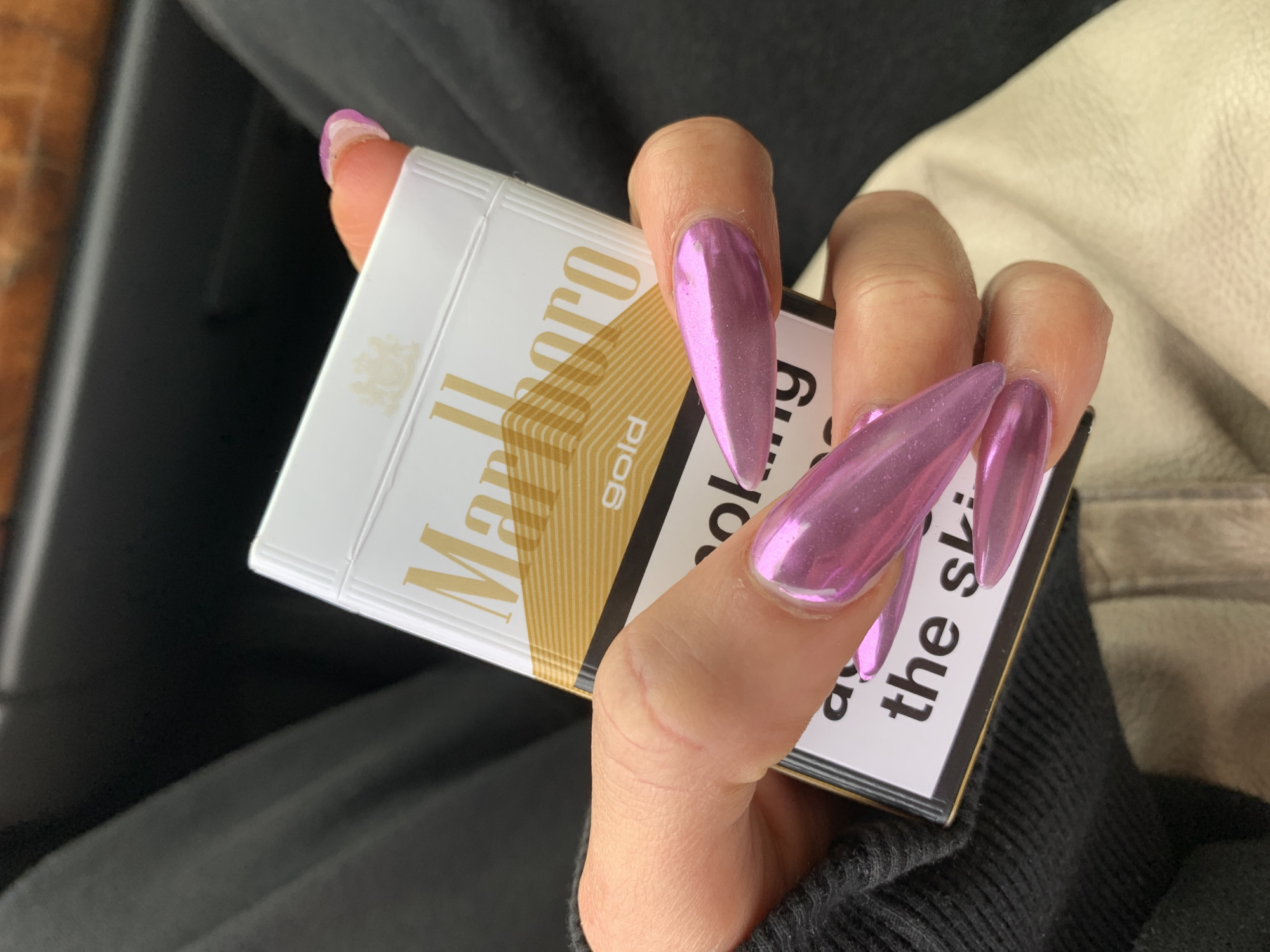 A close-up image features a hand holding a pack of Marlboro Gold cigarettes. The hand has long, sharply pointed (stiletto style) artificial nails with a distinctive metallic pink or light purple chrome finish. Minor chipping is visible on the nail of the pinky finger.
The cigarette pack is predominantly white and gold, clearly showing "Marlboro gold" branding and a subtle crest design above the brand name. A health warning label is partially visible on the side of the pack, with fragmented text reading "...ol...g" and "...the sl...".
The background is softly blurred, showing different textures. On the lower left, there is a dark, possibly ribbed fabric, suggesting clothing like a sweater sleeve. On the upper right, a lighter, smooth, off-white or beige material is visible, resembling upholstery such as a car seat or a jacket.
The scene appears to be an interior space, likely casual. No specific visual cues like time of day or identifiable landmarks are present within the image itself to confirm the provided location context of London, United Kingdom. The image captures the static action of holding the cigarette pack, with no discernible activity beyond that. No emotions are conveyed.