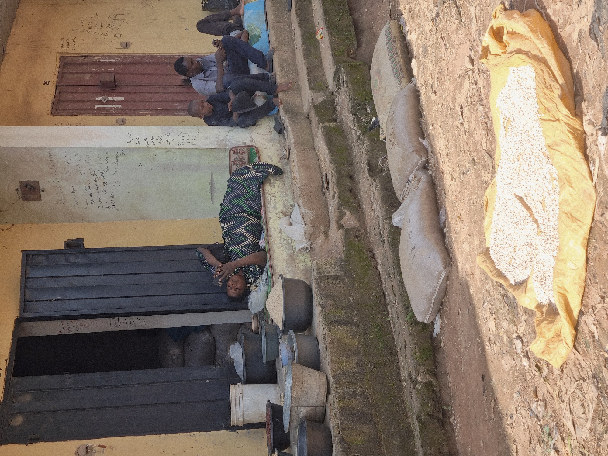 This image depicts an outdoor scene in New Nyanya, Nigeria. In the foreground, on the dirt ground, a yellow tarp is spread out, with a pile of small, pale grains on top. Behind this, on a tiered concrete structure, are several large sacks. Further back, against a yellow building with a dark metal door, a woman is lying on her side on a mat, looking at her phone. Beside her, on the concrete steps, sit two men. One man, appearing to be a child, is dressed in black and holding a black cap. The other man, wearing a light-colored short-sleeved shirt and dark pants, is also looking at his phone. More dark metal doors and a red-painted door are visible on the building, which has some chalked writing on its exterior wall. Various buckets and bowls, some containing grains or dark substances, are also present on the steps. The environment appears to be somewhat rustic, with some overgrowth on the concrete steps.