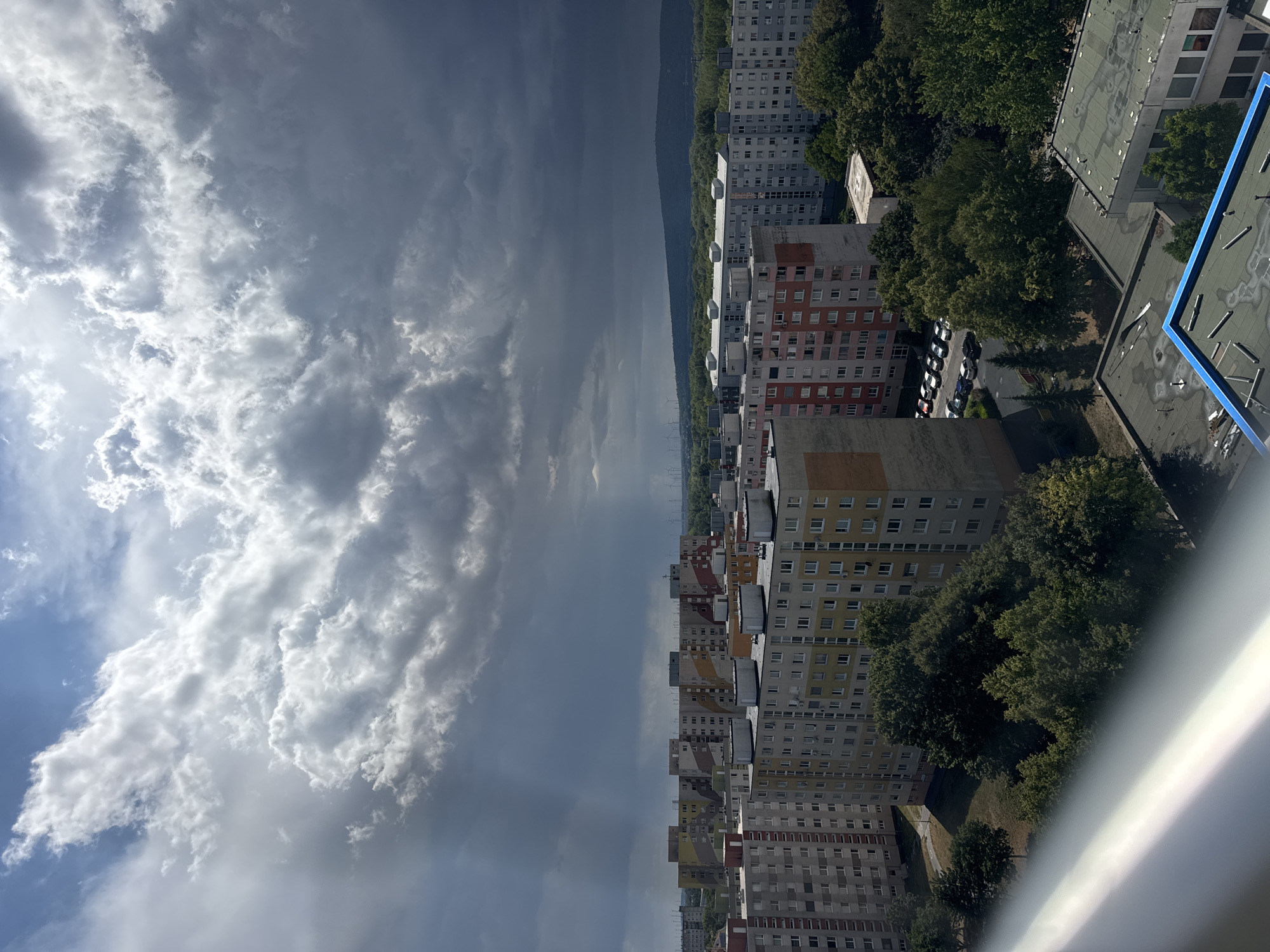 The image captures a panoramic view of the Petržalka district in Bratislava, Slovakia, under a dramatic sky. The foreground is dominated by a metallic railing, likely from a balcony or viewpoint, reflecting light. Beyond this, a dense cluster of identical, multi-story apartment buildings, typical of Soviet-era architecture, stretches across the frame. These buildings are characterized by their uniformly rectangular shapes, numerous windows, and varying color accents, including beige, yellow, and reddish-brown. Some buildings feature prominent roof structures.
In the middle ground, lush green trees form a dense canopy, interspersed with the apartment blocks and pathways. A parking lot filled with parked cars is visible between some of the buildings, indicating a residential area.
The background reveals a distant treeline and rolling hills, with a cluster of wind turbines visible on the horizon. The sky is a striking contrast, with bright sunlight breaking through dark, heavy clouds. Rays of light, often referred to as crepuscular rays, are visibly piercing through gaps in the clouds, illuminating parts of the cityscape and the sky. The overall impression is one of a dynamic weather event unfolding over a typical urban residential landscape.