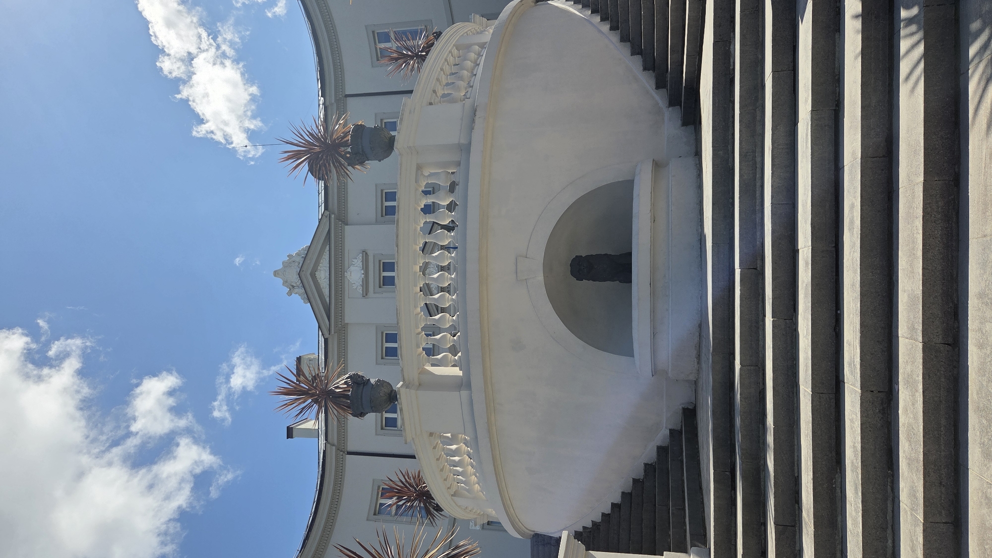 The image shows a wide, curved white stone staircase leading up to a semicircular building. The staircase is bordered by grey stone steps and has a white balustrade along its upper section. At the top of the stairs, on a circular terrace, are two large planters with spiky, reddish-brown plants. In a semicircular niche below the balustrade is a dark statue, possibly of a dog. The building behind the terrace is light-colored with multiple windows. The sky above is bright blue with scattered white clouds. This scene is located at the Palanga Amber Museum in Palanga, Lithuania.