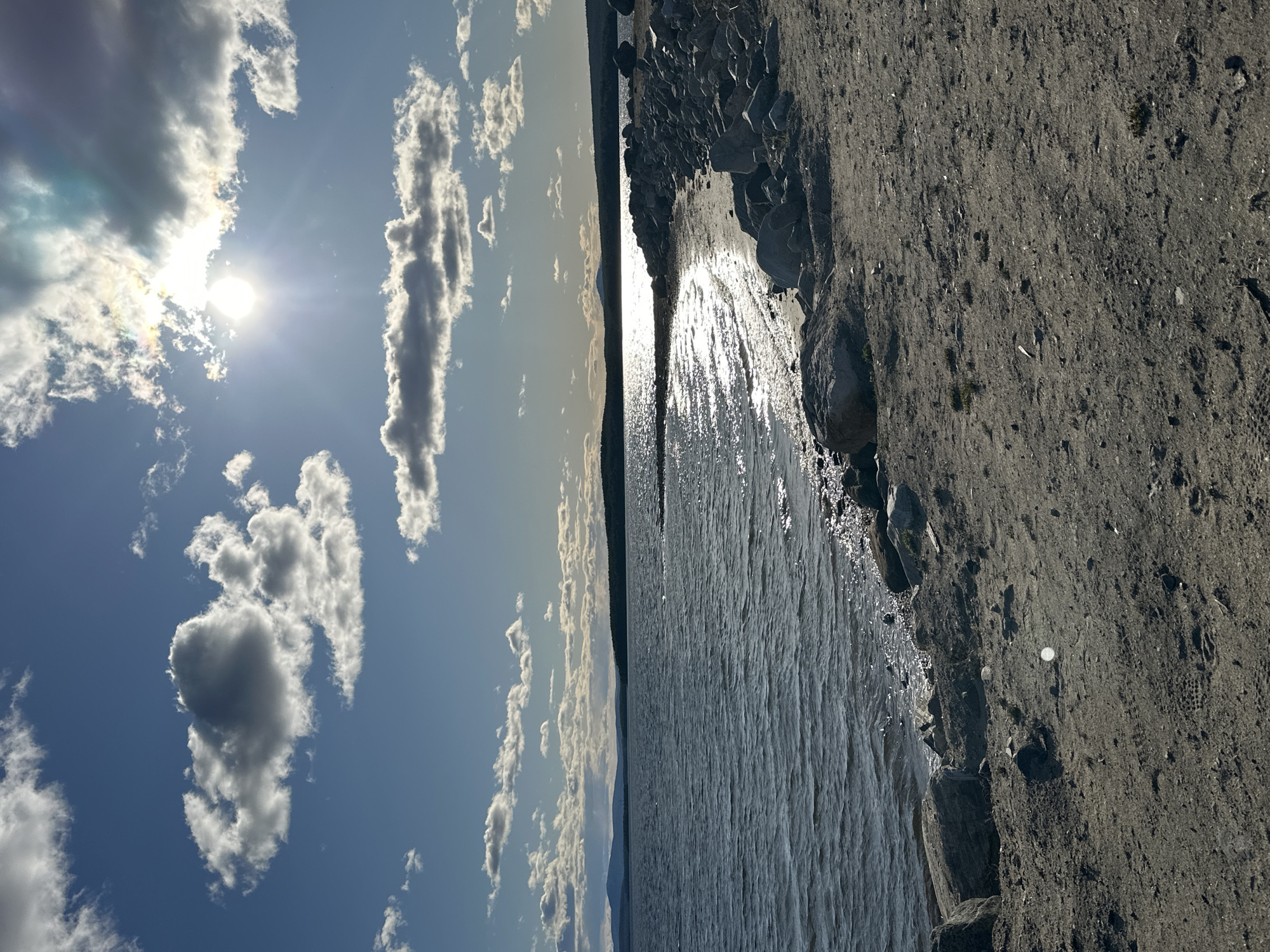 The image depicts a body of water, possibly a lake or sea, meeting a rocky shoreline under a sky with scattered clouds and a visible sun. The ground alongside the water appears to be made of dark-colored sediment or soil interspersed with rocks. The sun's reflection glints off the water's surface. The location is unknown.