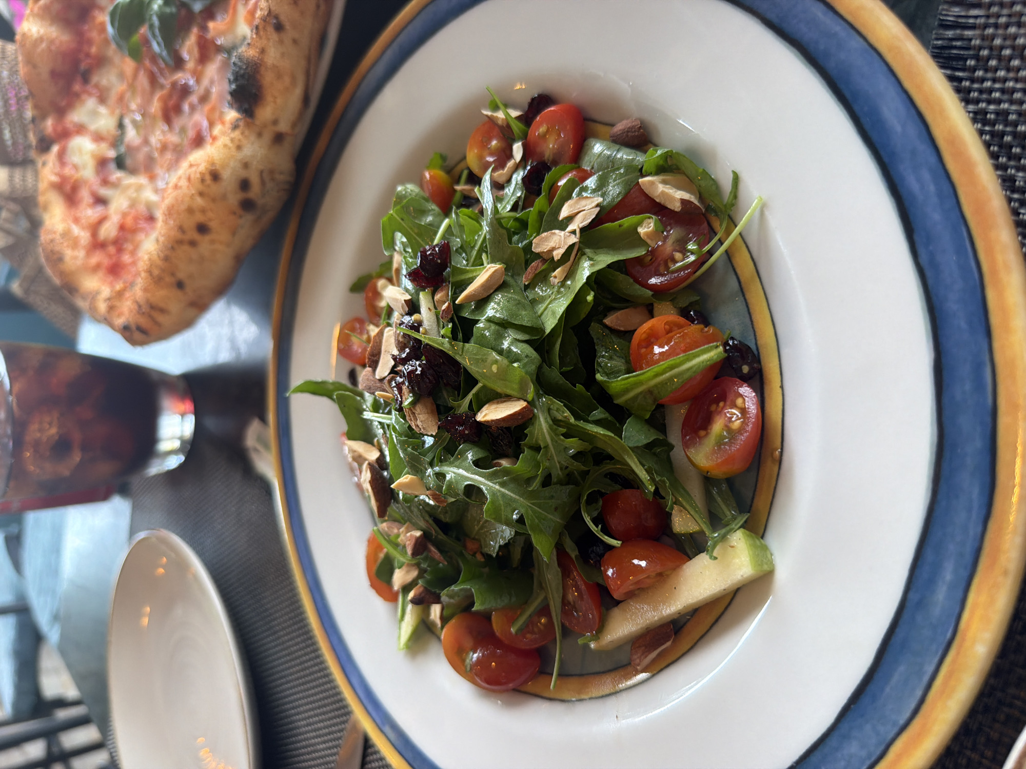 An overhead shot captures a vibrant salad on a white plate with a decorative blue and yellow rim. The salad is a medley of fresh green arugula, halved cherry tomatoes, dried cranberries, and slivered almonds. A pale green sliver, possibly apple or pear, is also visible. In the background, a blurry pizza with melted cheese and basil rests on a dark surface. A glass of dark liquid, likely soda or iced tea, and a white plate are also present. The setting appears to be a restaurant with a muted, textured tablecloth. The scene is set in Baan Bo Phut, Thailand.