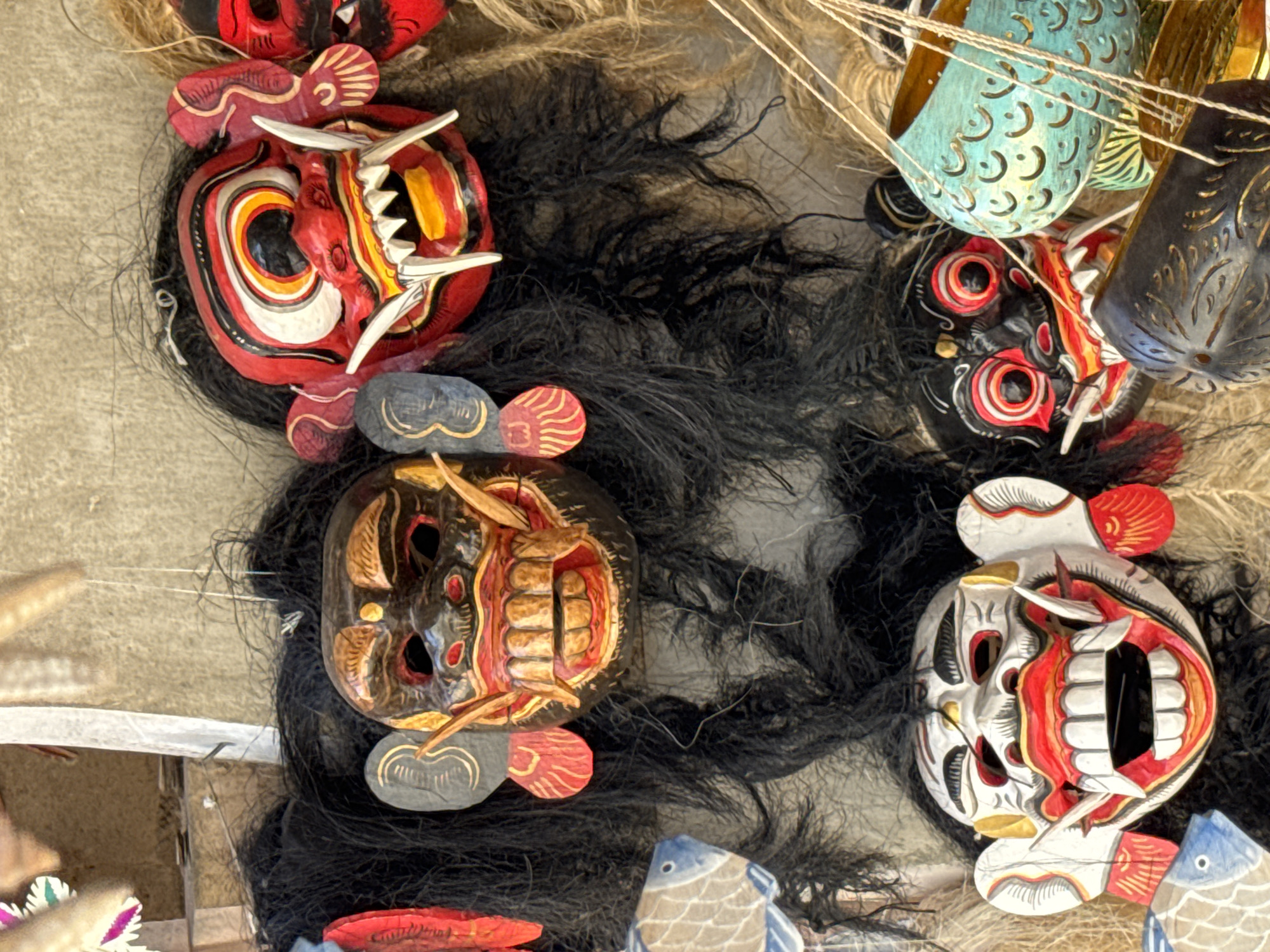 In Ubud District, Indonesia, a collection of Balinese masks with elaborate headdresses and string decorations are displayed against a textured wall. The masks feature intricate carvings, painted details in red, white, yellow, and black, and exaggerated facial expressions with sharp teeth. Some masks have black, shaggy hair extensions, while others are adorned with smaller carved ornaments and string. To the left, a decorative blue and white fish sculpture is partially visible. In the lower right, a greenish-blue carved object, possibly a bowl or lantern, hangs with thin strings. The overall scene suggests a market or artisan display of traditional Indonesian crafts.