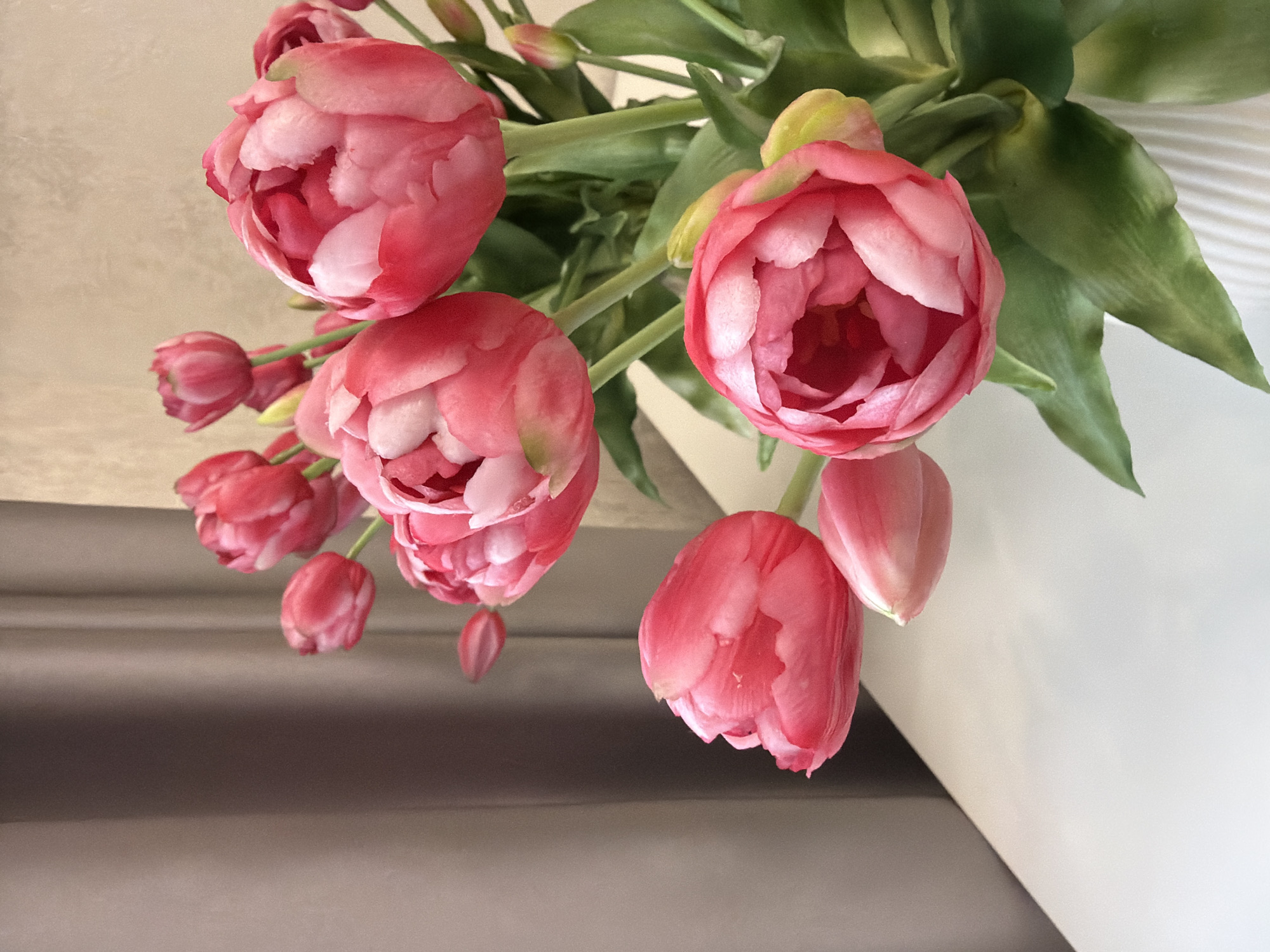 A bouquet of pink double tulips is displayed in a white ribbed vase on a white surface. The tulips are a mix of fully bloomed and partially opened flowers, with some buds visible. The background consists of a neutral-toned curtain on the left and a textured wall on the right. The lighting is soft, creating a gentle illumination of the flowers.