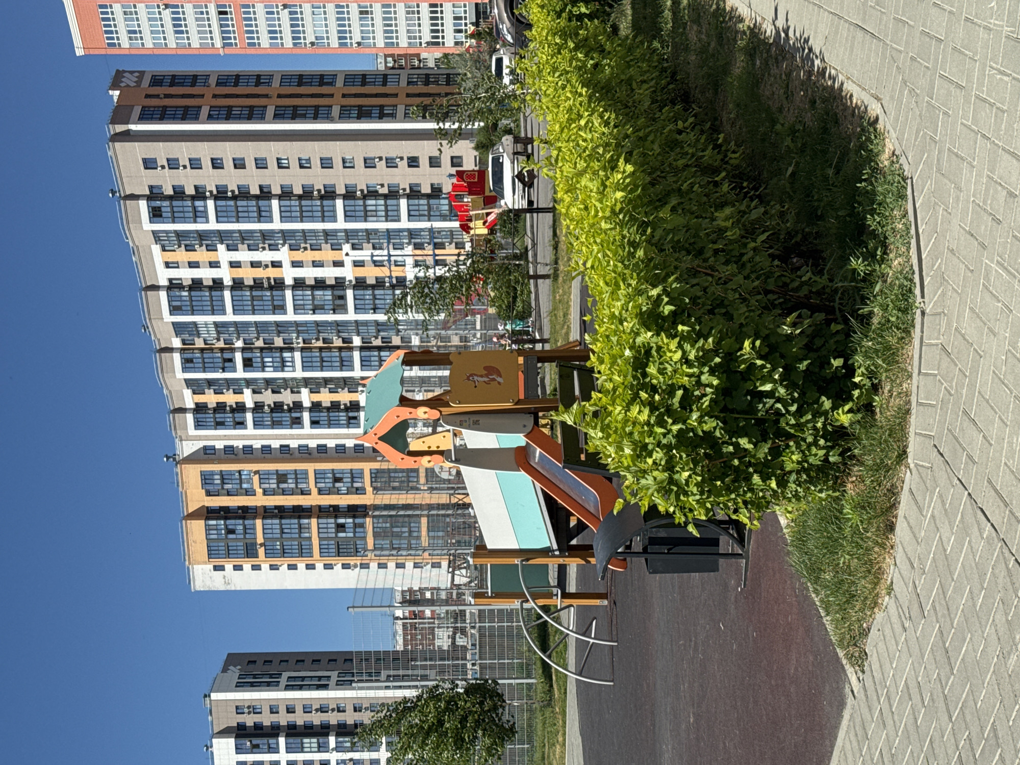 The image shows a playground in a residential area with several multi-story apartment buildings in the background. The playground features a slide and climbing structure, a metal arch climber, and a trash bin. A well-maintained green hedge runs along the edge of the playground, bordering a brick-paved walkway. Several cars are parked on the street in front of the buildings, and a few people are visible in the distance. The sky is clear and blue, indicating a sunny day. The architectural style of the buildings suggests a modern urban development. The presence of a playground and apartment buildings indicates a residential area likely designed for families. The overall scene depicts a typical day in a modern residential neighborhood in Barnaul, Russia, characterized by new construction and organized public spaces.