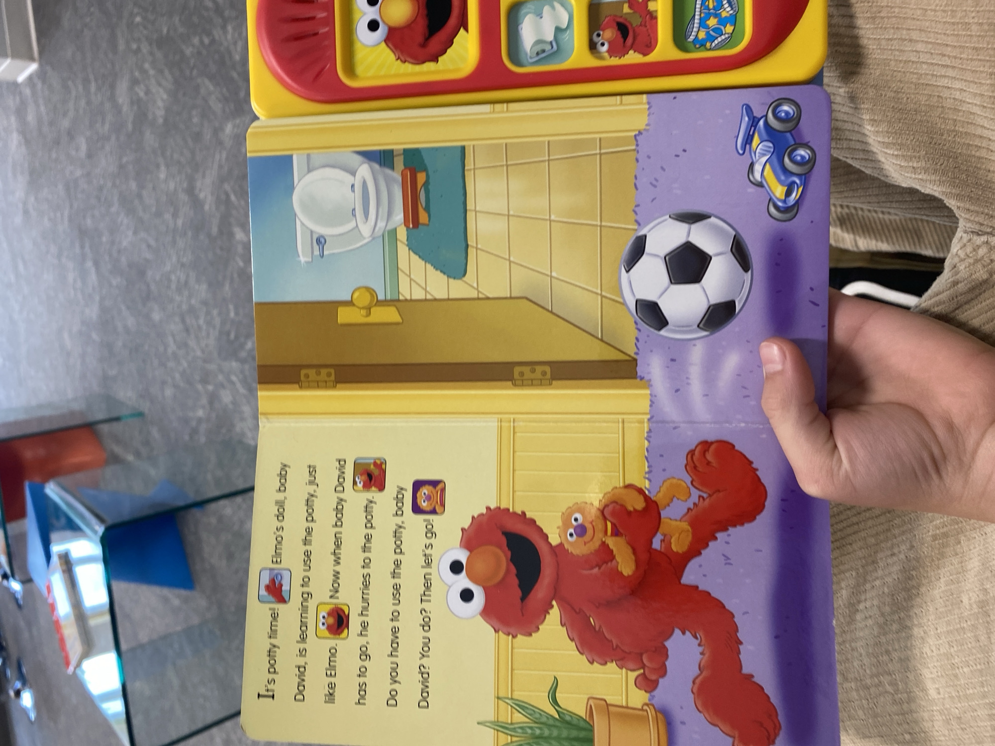 The image shows an open children's book on a patterned floor. The left page has text and illustrations related to potty training, featuring characters Elmo and baby David. Elmo is depicted holding a baby, running. The right page is a part of an interactive toy, with buttons and images of Elmo. In the foreground, a person's hand is visible, holding the book open. The background shows a blurred interior, possibly a living room with glass furniture. The scene appears to be indoors, with no indication of its specific location in Chesapeake, United States of America.