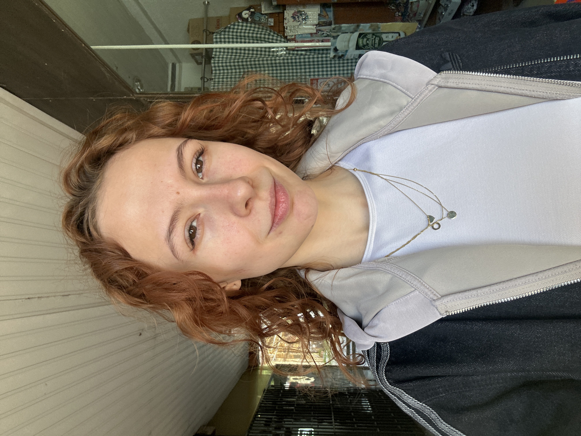 A woman with curly, reddish-brown hair is centered in the image, positioned in front of a building with a weathered, pale wooden facade. She is wearing a white t-shirt, a light gray zip-up hoodie, and a dark-colored jacket. She wears a delicate, layered necklace with small pendant charms. The background behind her consists of a shop window with a reflection of various objects, including a green and white checkered pattern item and some boxes. Below the shop window, there is a gated area with metallic bars. Given the architectural style and the appearance of the surrounding environment, it's likely this photograph was taken in a street or public area in Novi Sad, Serbia.