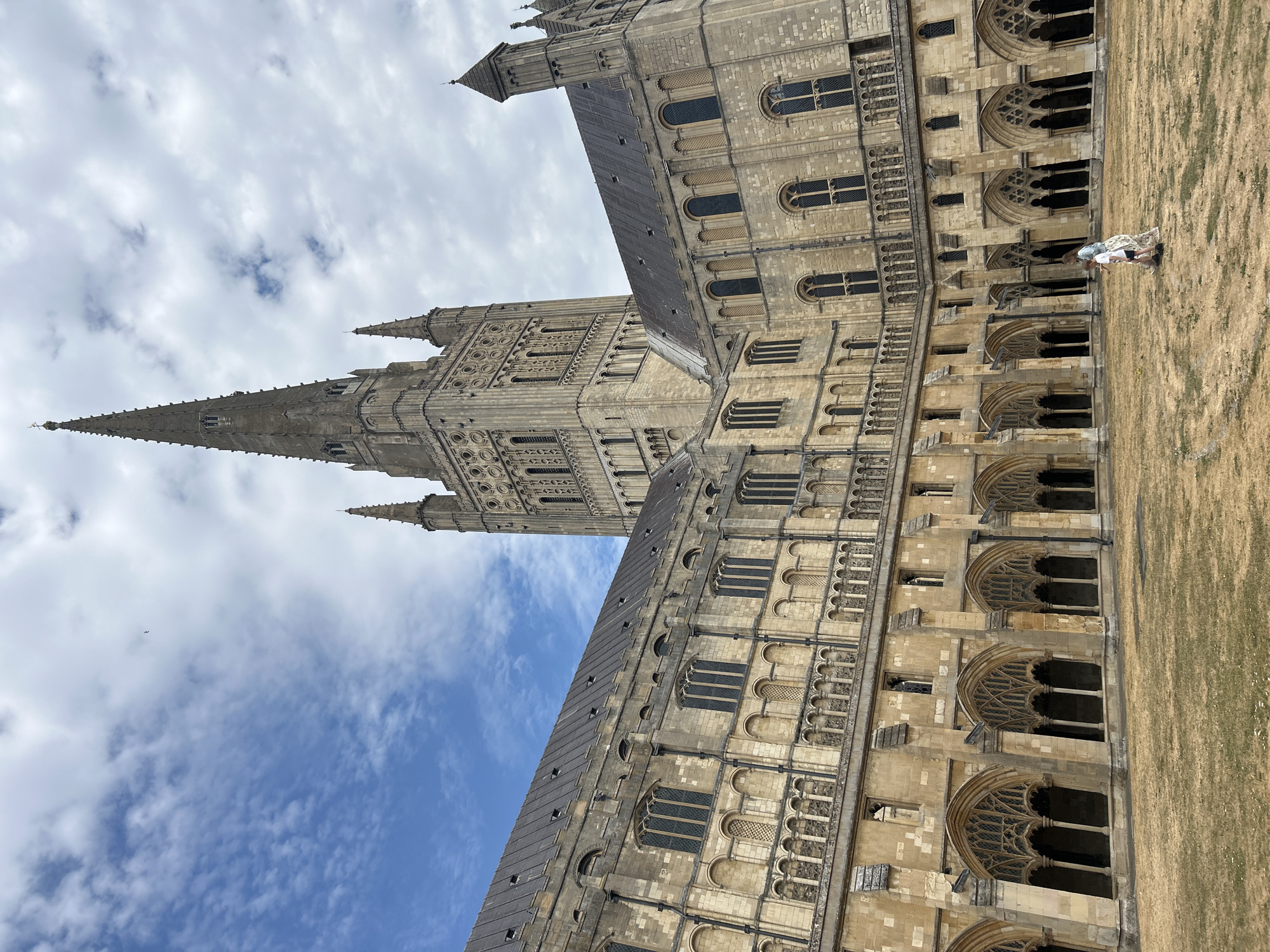 The image shows a view of the Norwich Cathedral in Norwich, United Kingdom. The cathedral is a large, historic building with a tall spire. The foreground of the image is a courtyard with dry grass. Two people, a woman and a child, are walking across the courtyard. The sky is partly cloudy with blue sky visible. The architecture of the cathedral is gothic, with arched windows and intricate stone carvings. The lighting suggests it is daytime, possibly mid-morning or afternoon.