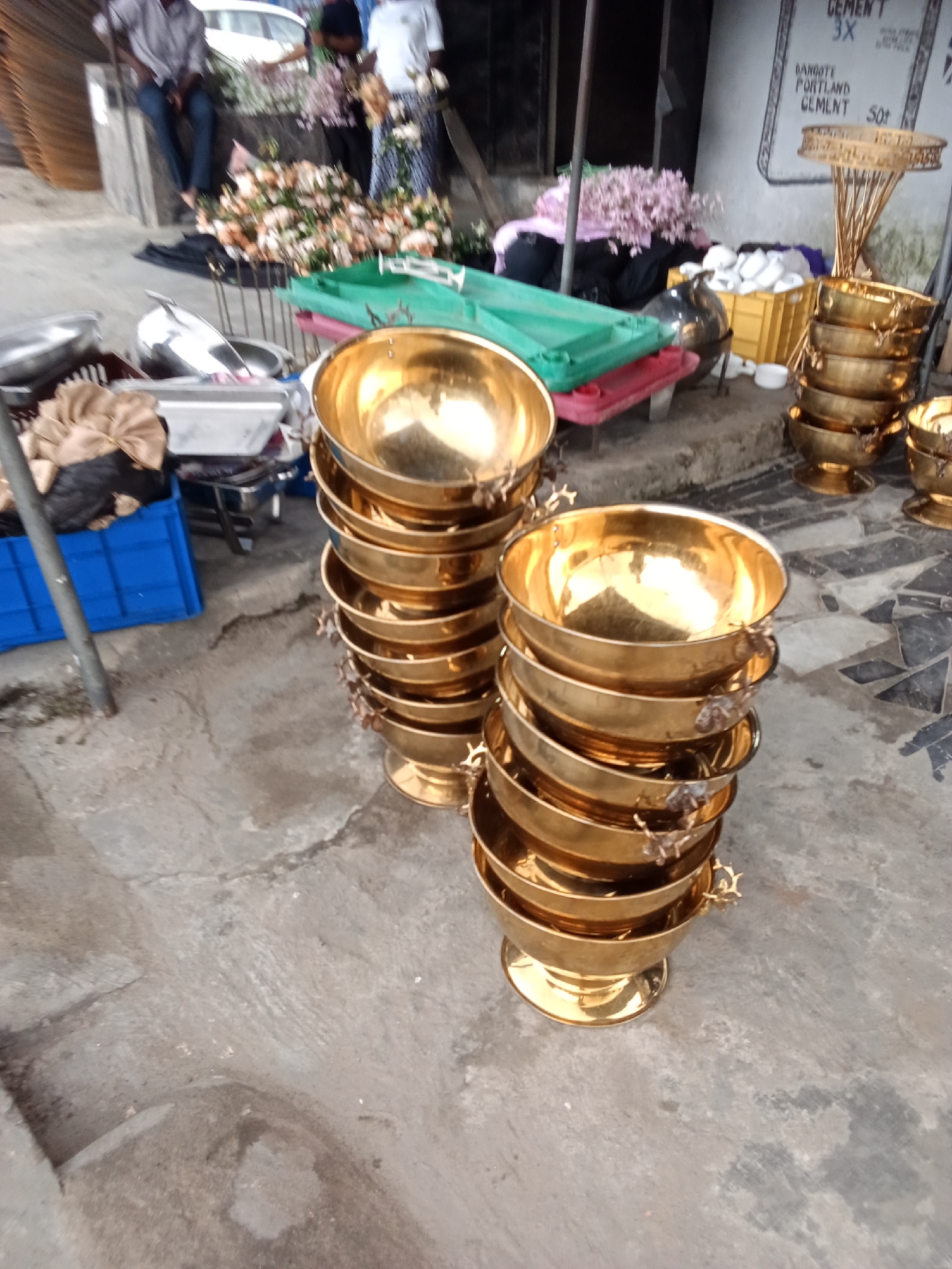 The image shows an outdoor scene in Ado, Nigeria. Two stacks of approximately ten gold-colored bowls with handles are positioned on a concrete surface. In the background, there are people standing, a flower arrangement, containers, and miscellaneous objects. A sign reading "CEMENT" is visible on a wall.
