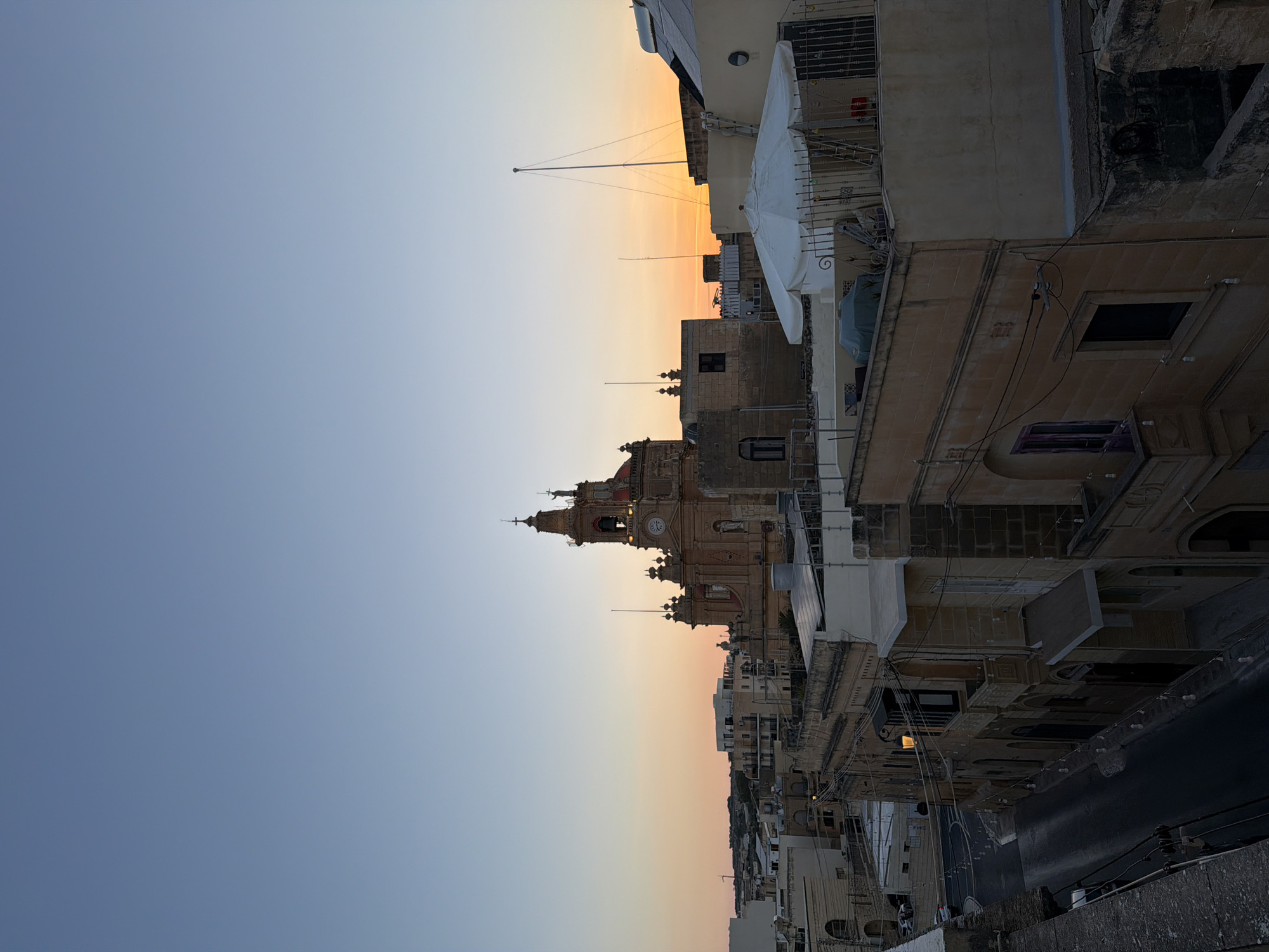 The image depicts an elevated view of the city of Fontana, Malta, at dusk. A prominent church with a clock tower stands in the center, its dome and bell tower illuminated by the fading light. The sky transitions from a clear blue at the top to a warm orange and pink hue near the horizon, indicating sunset. Below the church, a dense collection of buildings with terracotta roofs and stone facades lines narrow streets. Some buildings have balconies and arched windows, typical of Maltese architecture. A white umbrella is visible on a rooftop in the foreground, along with a ladder and some construction materials. The street below is dimly lit, with streetlights casting a soft glow. The scene is tranquil, capturing the quiet beauty of the city as day turns into night.
