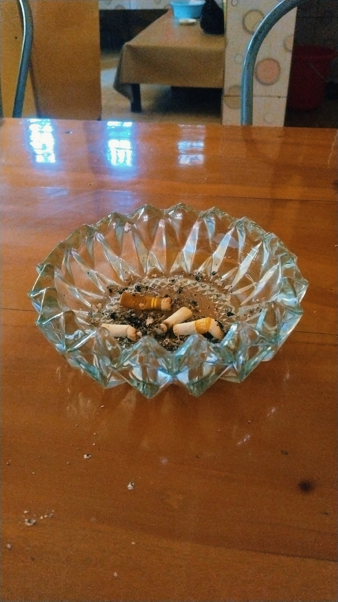 The image shows a glass ashtray filled with cigarette butts and ash, placed on a wooden table. In the background, a tablecloth-covered table and chairs are visible, suggesting an indoor setting. The lighting indicates it might be daytime with light filtering in from windows, creating reflections on the table surface. The scene appears to be a common area, possibly a dining room or cafe, in a private residence or establishment. The presence of an ashtray implies that smoking has occurred recently in this location. The image does not contain any specific identifiers that would place it in Besuki, Indonesia.