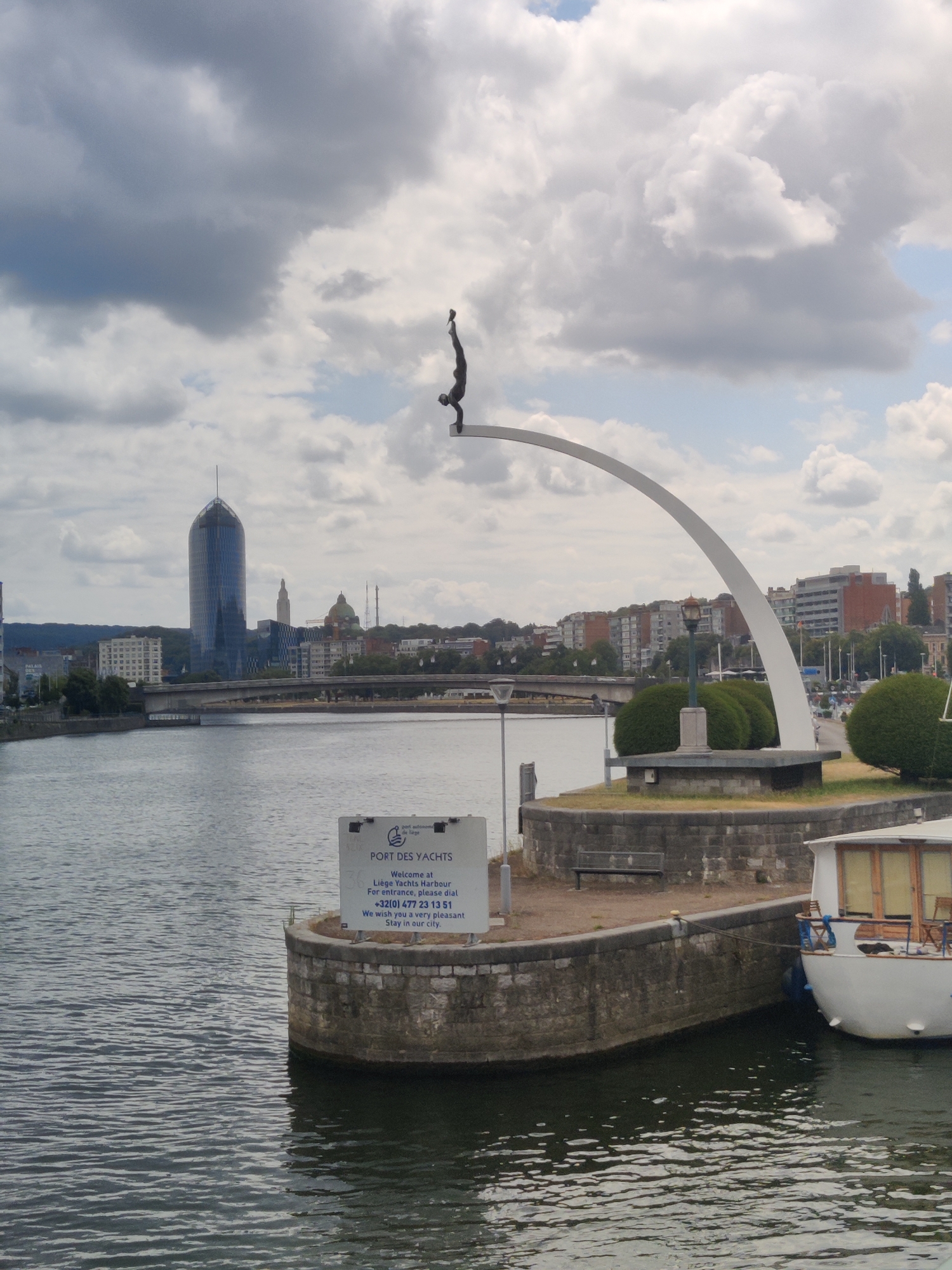 The image captures a view of the River Meuse in Liège, Belgium, with a prominent sculpture of a diver in mid-air. The sculpture, a bronze figure in a diving pose, is perched atop a white arched structure that extends over the water. In the background, the modern skyscraper of the Liège-Guillemins railway station dominates the skyline, its glass facade reflecting the cloudy sky. The cityscape also features other buildings, bridges, and greenery. In the foreground, a stone quay juts out into the river, adorned with a sign for "PORT DES YACHTS" and "Liège Yacht Harbour." A portion of a white boat is docked at the quay, its windows and wooden railings visible. The water of the river is calm, with gentle ripples reflecting the overcast sky. The scene suggests a typical day at the harbor, with a blend of modern architecture and recreational activity.