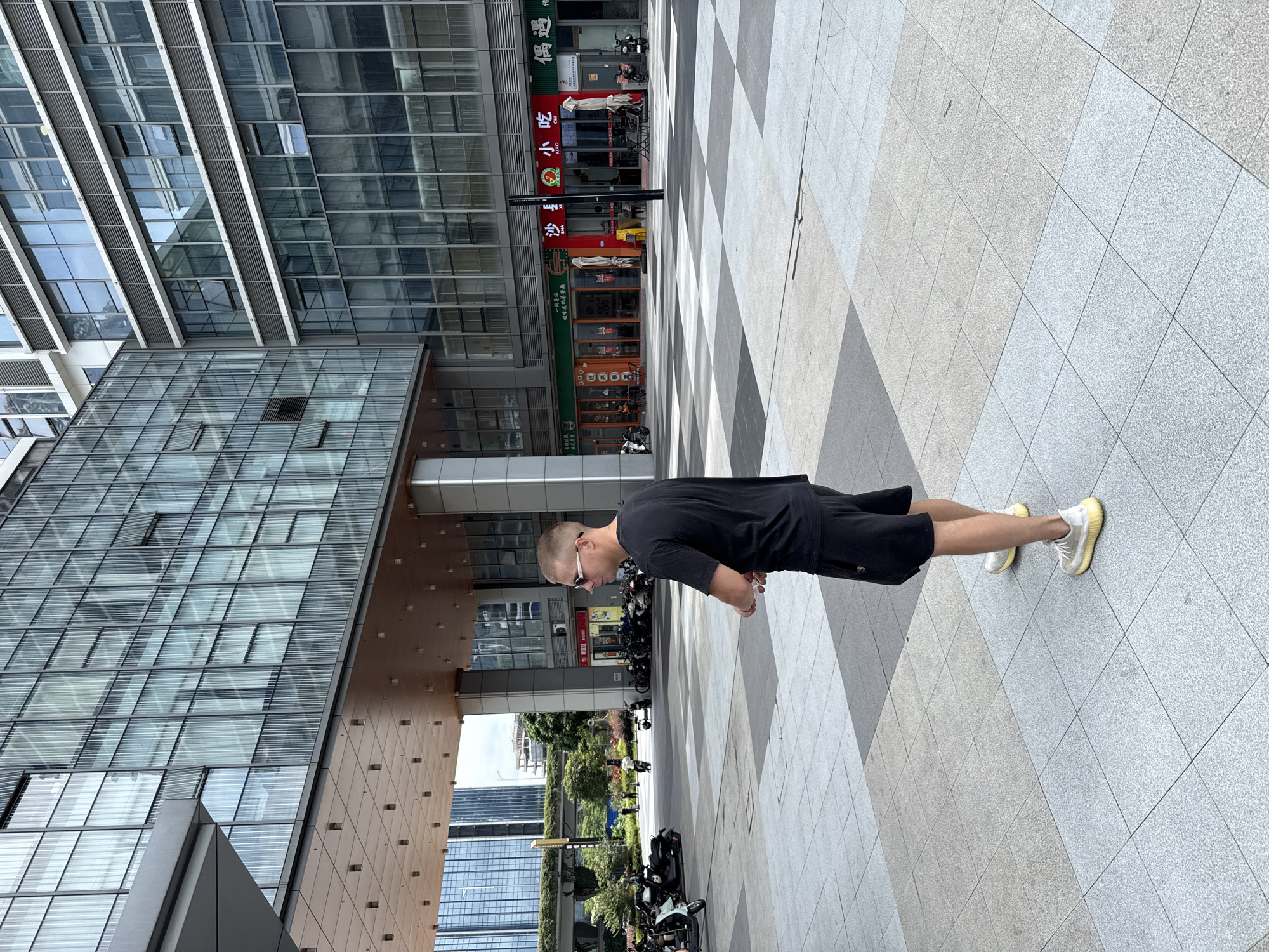 The image depicts an outdoor scene in the Huangpu District of China, likely within a modern commercial area. In the foreground, a young man with short hair and sunglasses is standing, facing away from the camera, wearing a black t-shirt and black shorts, with white and yellow sneakers. He is standing on a tiled plaza with a checkerboard pattern of light and dark gray tiles.
Behind the man, a multi-story building with a glass facade dominates the background. The building has numerous windows and architectural elements that suggest a contemporary design. Below the glass facade, there is a row of storefronts, some of which display signage with Chinese characters, indicating commercial establishments such as restaurants. Several motorcycles and scooters are parked in front of these storefronts and in the plaza area.
Further in the background, other modern buildings are visible, along with some greenery and trees. The sky appears overcast, suggesting a daytime scene. The overall impression is one of an urban environment with a mix of commercial and pedestrian spaces.